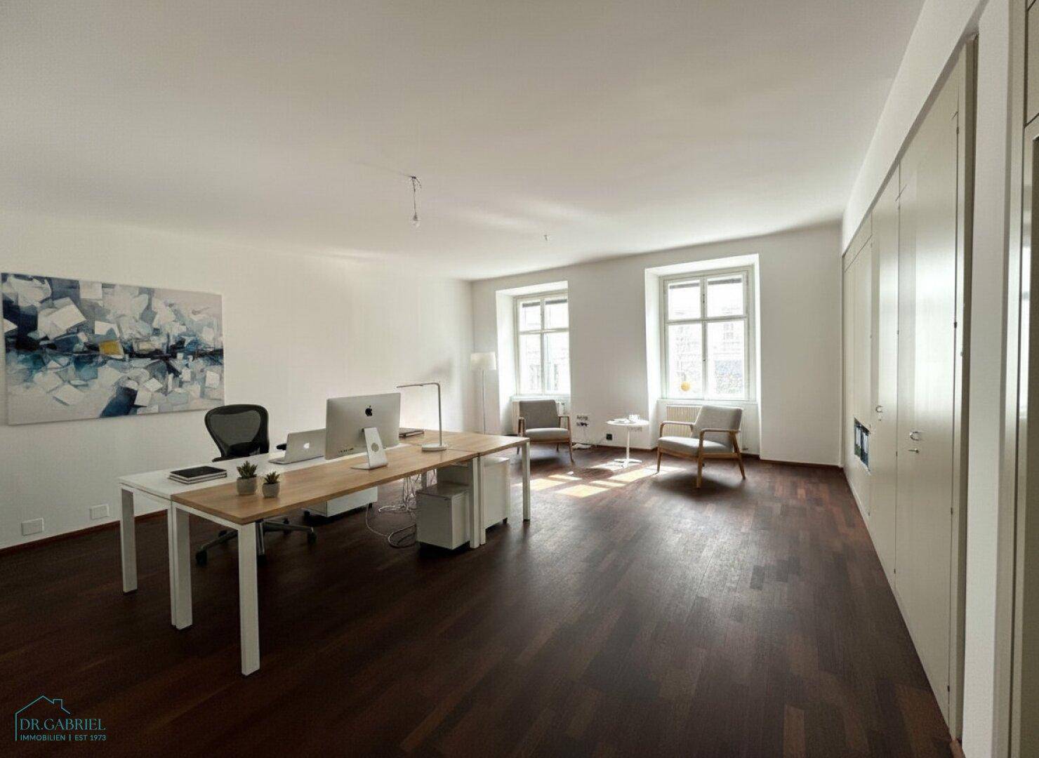 Geräumiges Büro mit dunklem Holzboden, weißen Wänden und modernen Arbeitsplätzen, gut beleuchtet durch große Fenster.