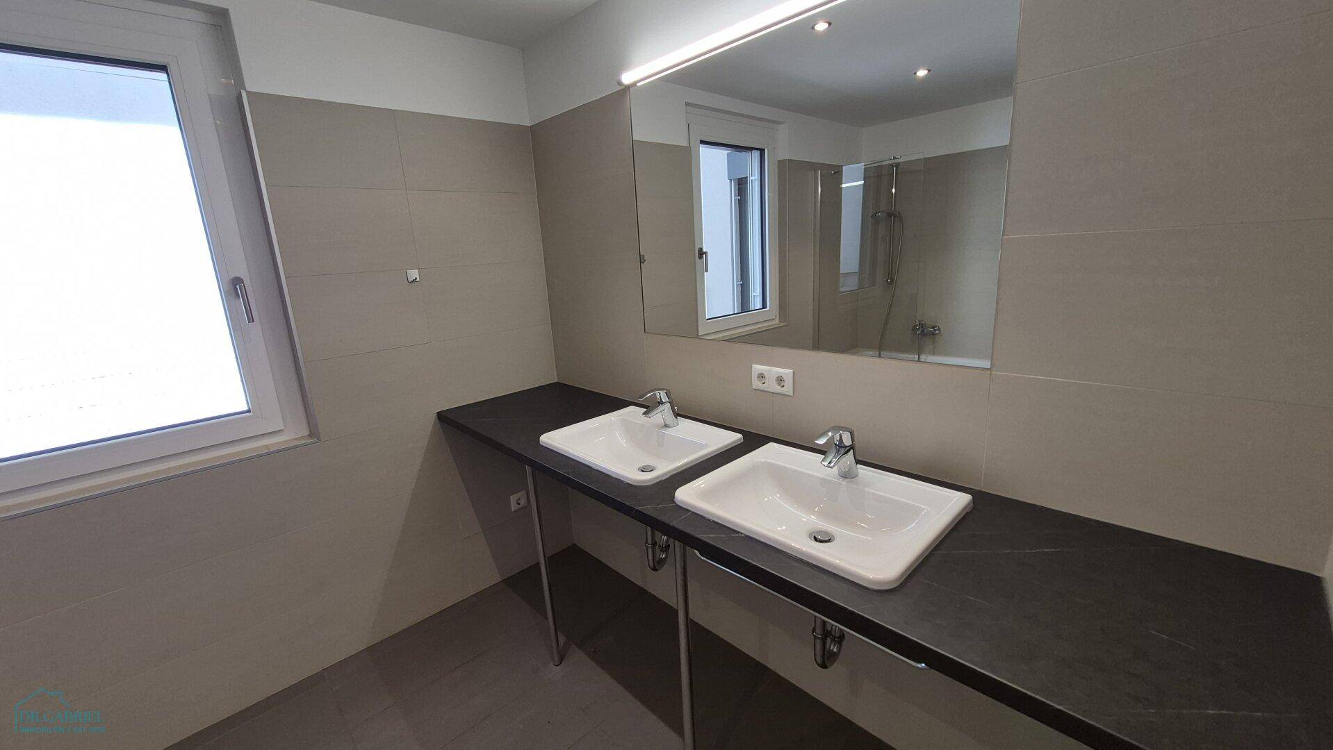 Helles Badezimmer mit Doppelwaschbecken, großem Spiegel und Fenster, modern gefliest.