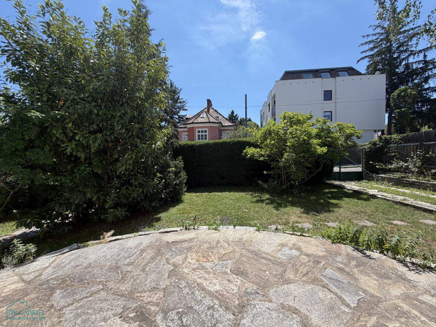 Gartenbereich mit Rasenfläche und gepflastertem Sitzplatz, umgeben von Hecken und Bäumen.