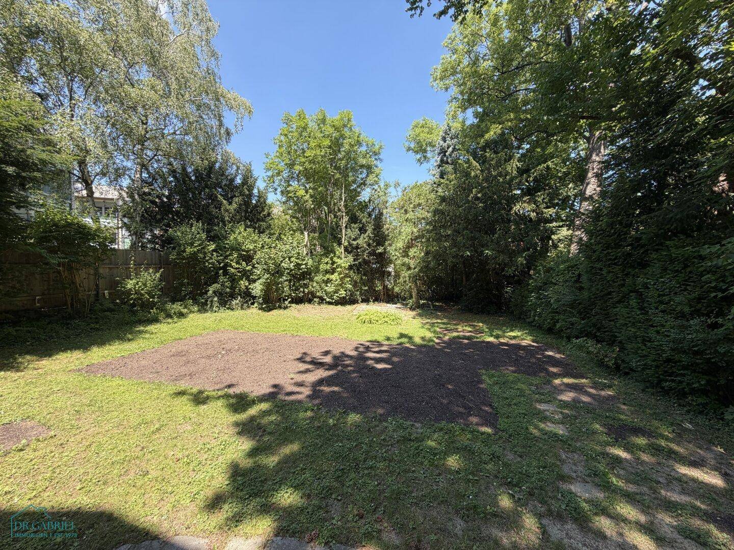 Großer, gepflegter Gartenbereich mit Rasenfläche und altem Baumbestand unter blauem Himmel.