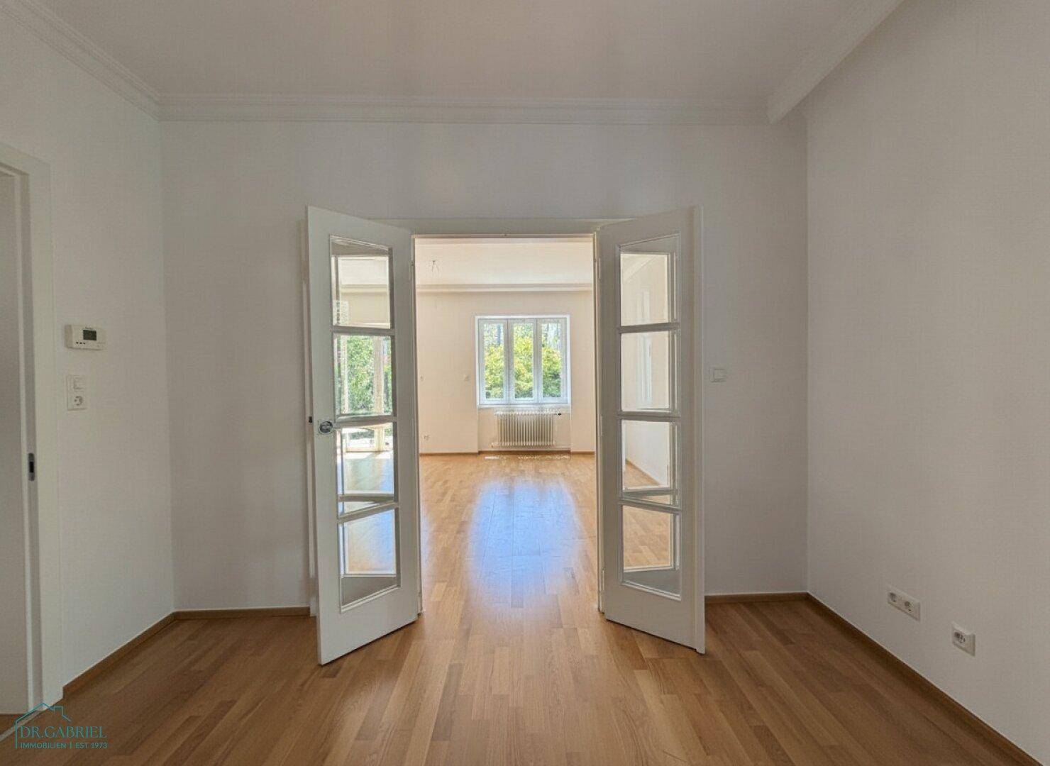 Durchgang zwischen zwei hellen Räumen mit Parkettboden und großen Fenstern.