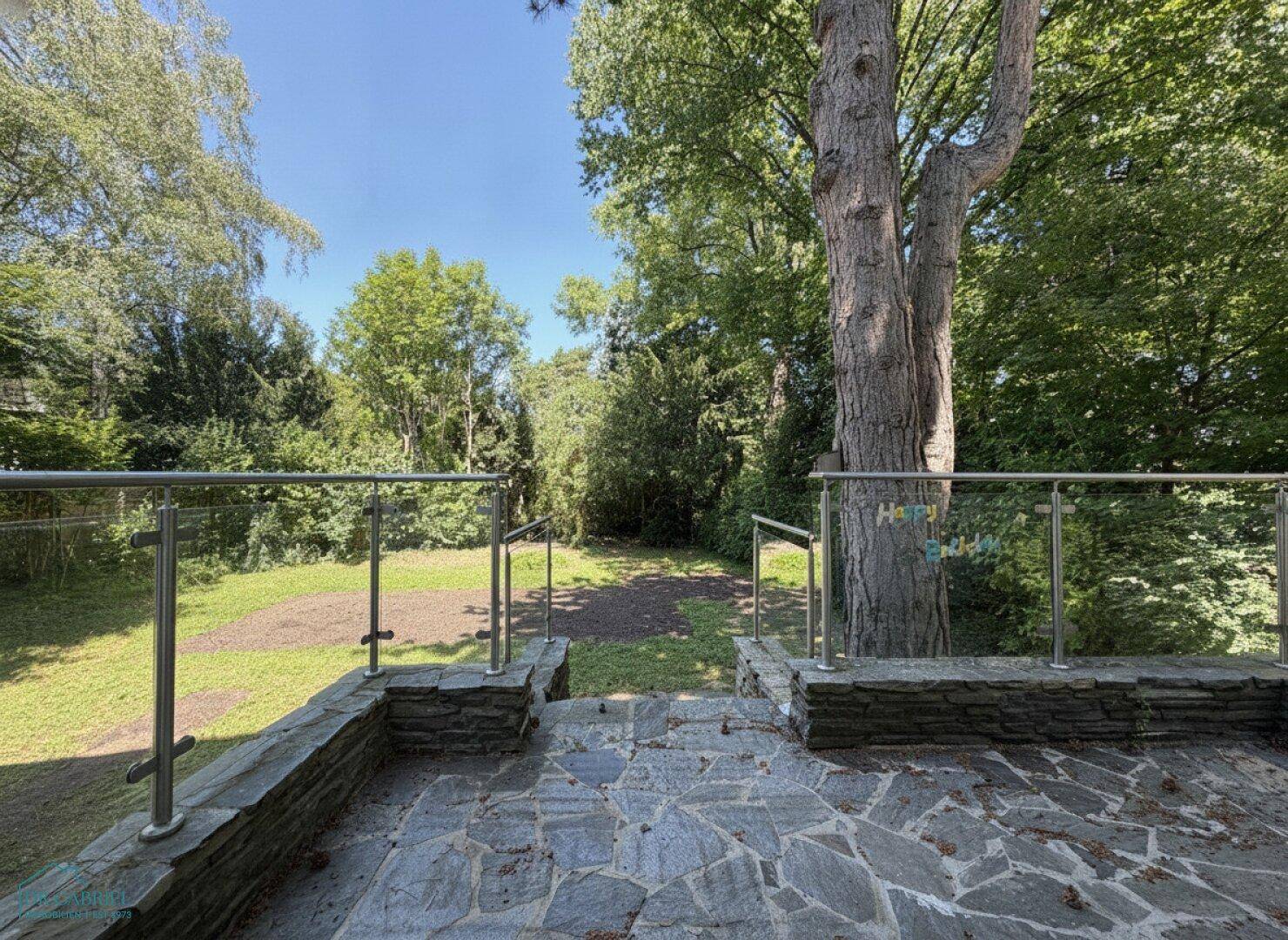 Gepflasterter Bereich mit Glasgeländer, der zu einem weitläufigen Garten mit altem Baumbestand führt.
