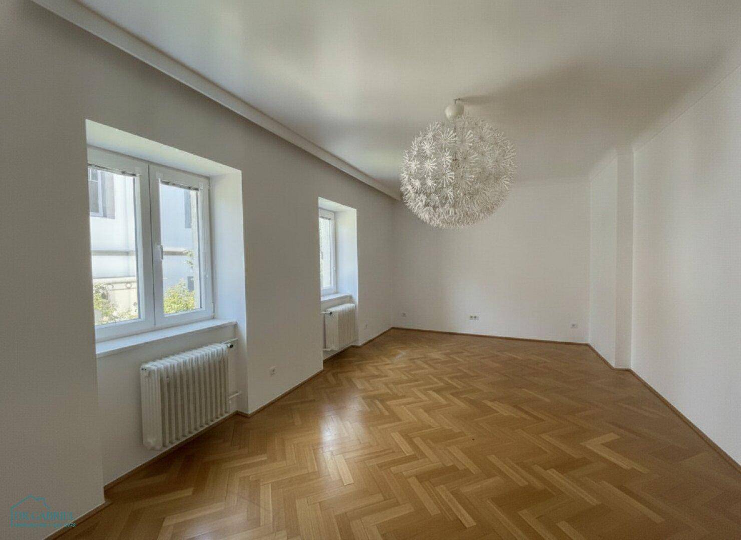 Geräumiges Wohnzimmer mit Fischgrätparkett, mehreren Fenstern und moderner Deckenleuchte.
