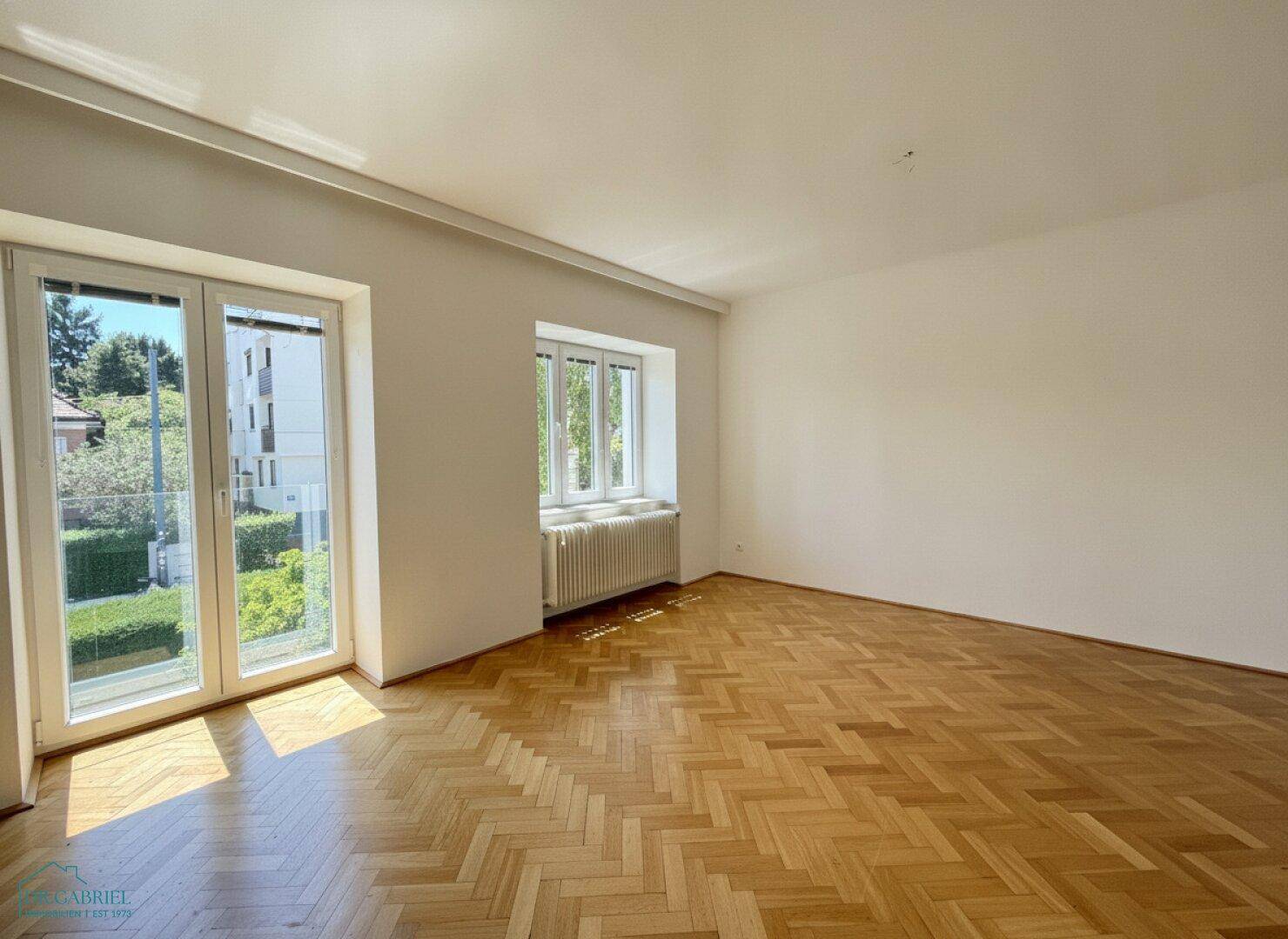 Helles Wohnzimmer mit Fischgrätparkett und Zugang zu einem Balkon mit Blick ins Grüne.