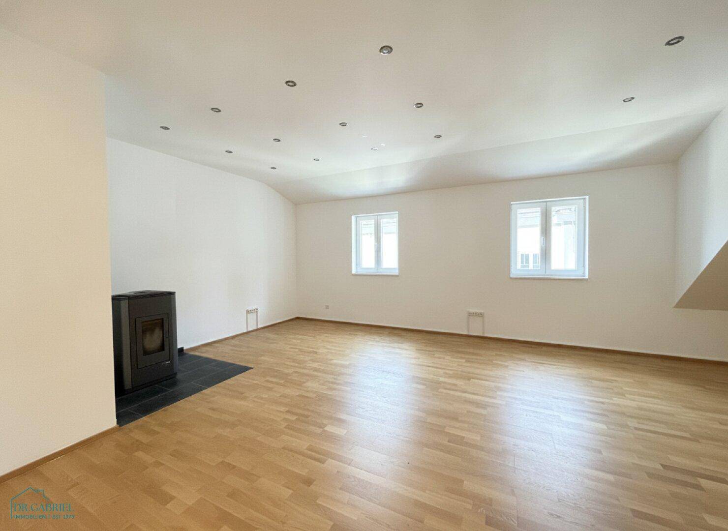 Geräumiger Dachgeschossraum mit Parkettboden, Kaminofen und mehreren Fenstern.