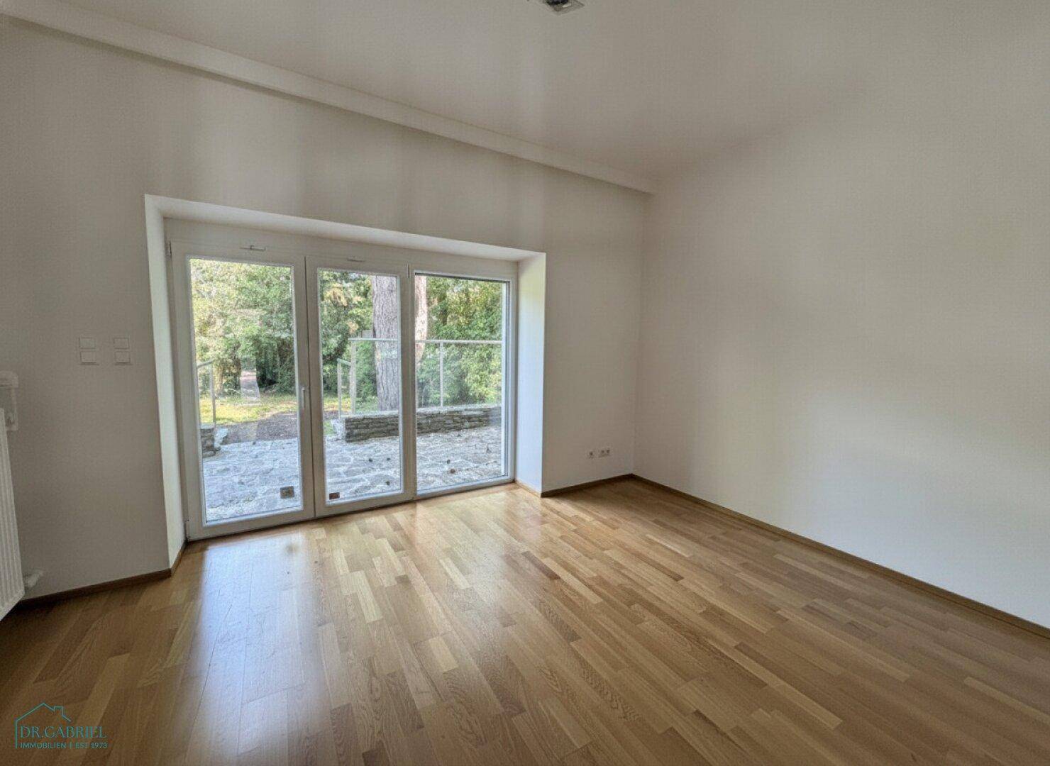 Heller Raum mit Parkettboden und großen Fenstertüren, die zum Gartenbereich führen.