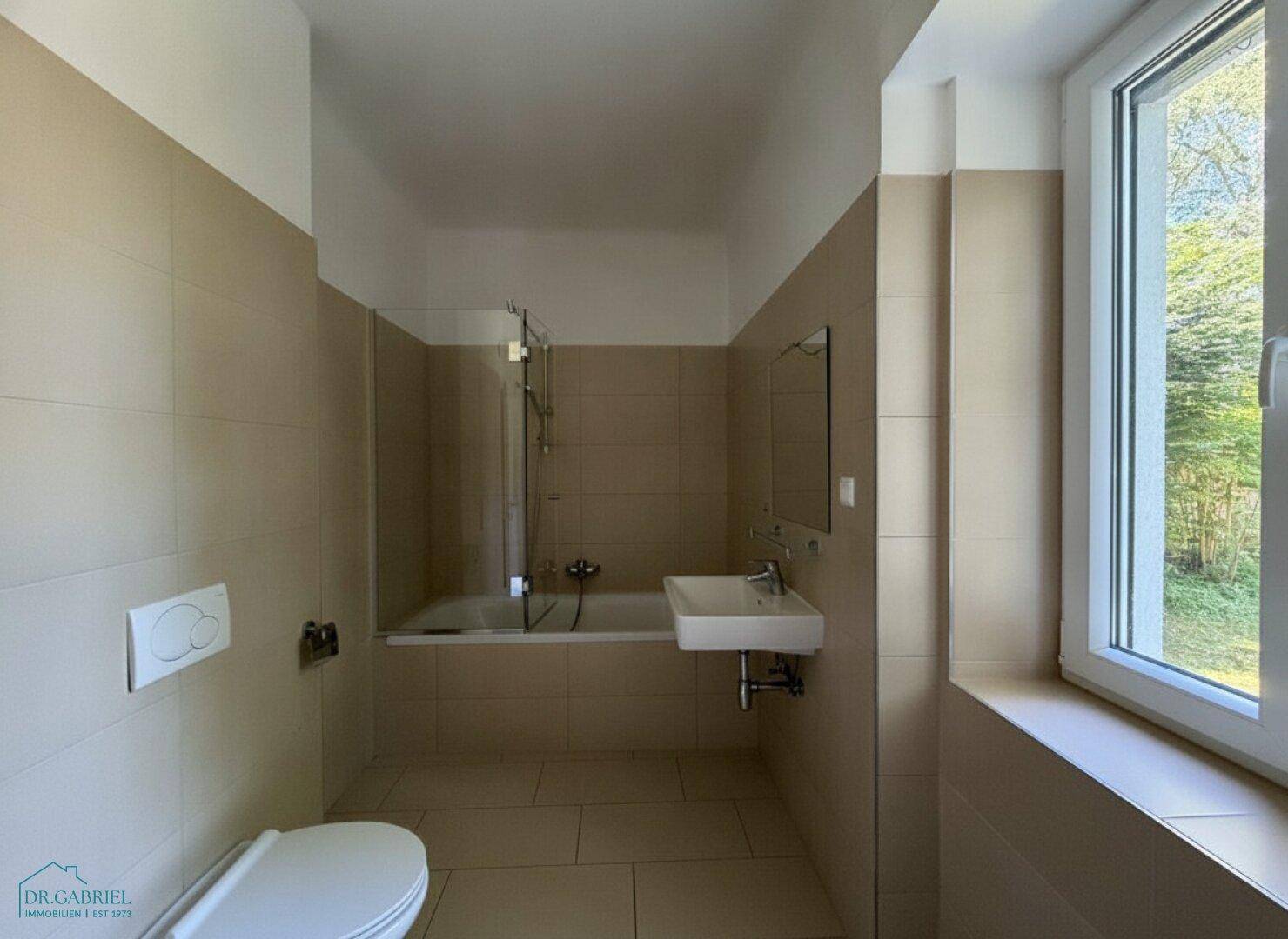 Gefliestes Badezimmer mit Badewanne, Waschbecken, Toilette und Fenster mit Blick ins Grüne.