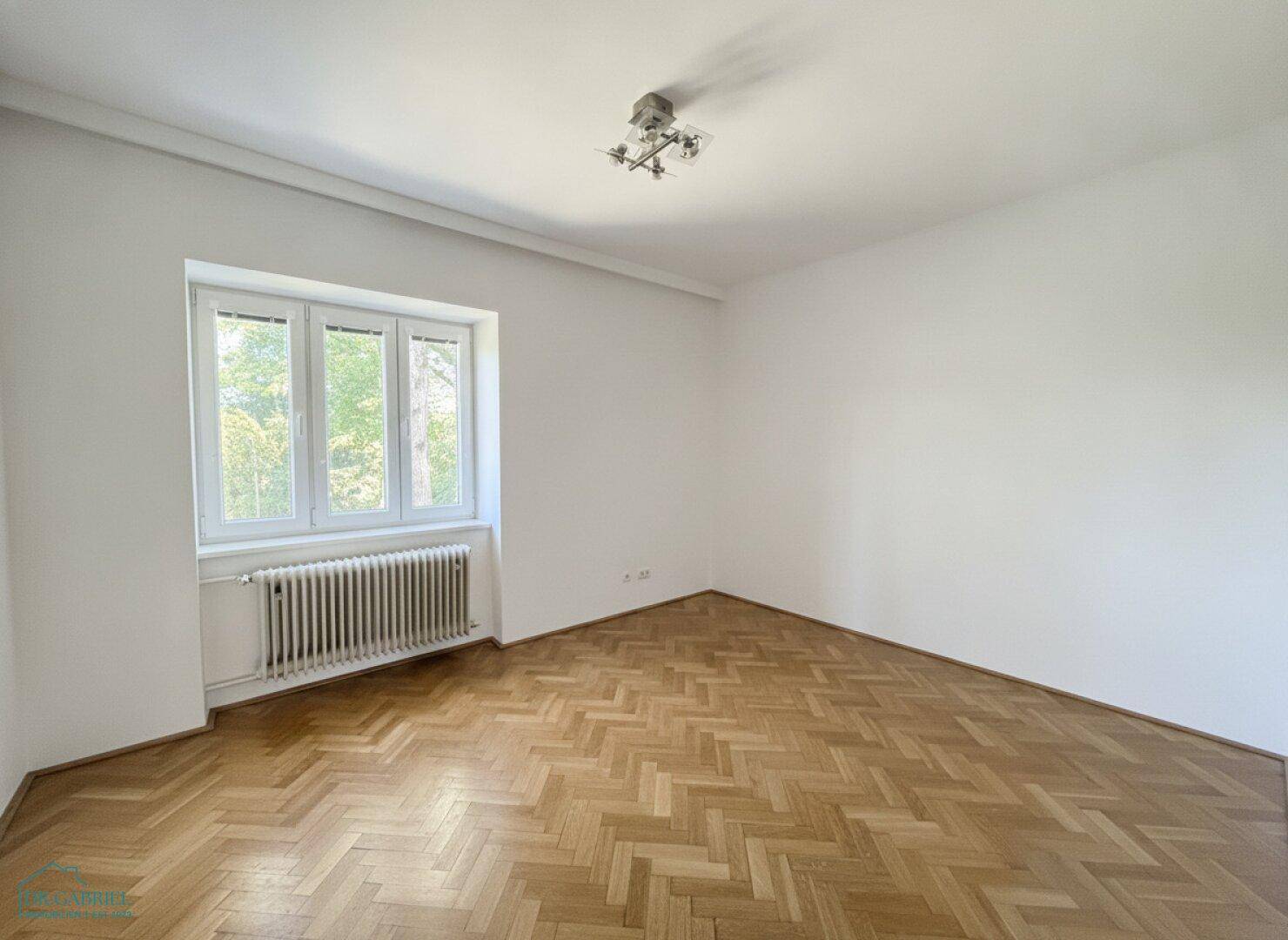Helles Schlafzimmer mit Fischgrätparkettboden und großem Fenster mit Blick ins Grüne.