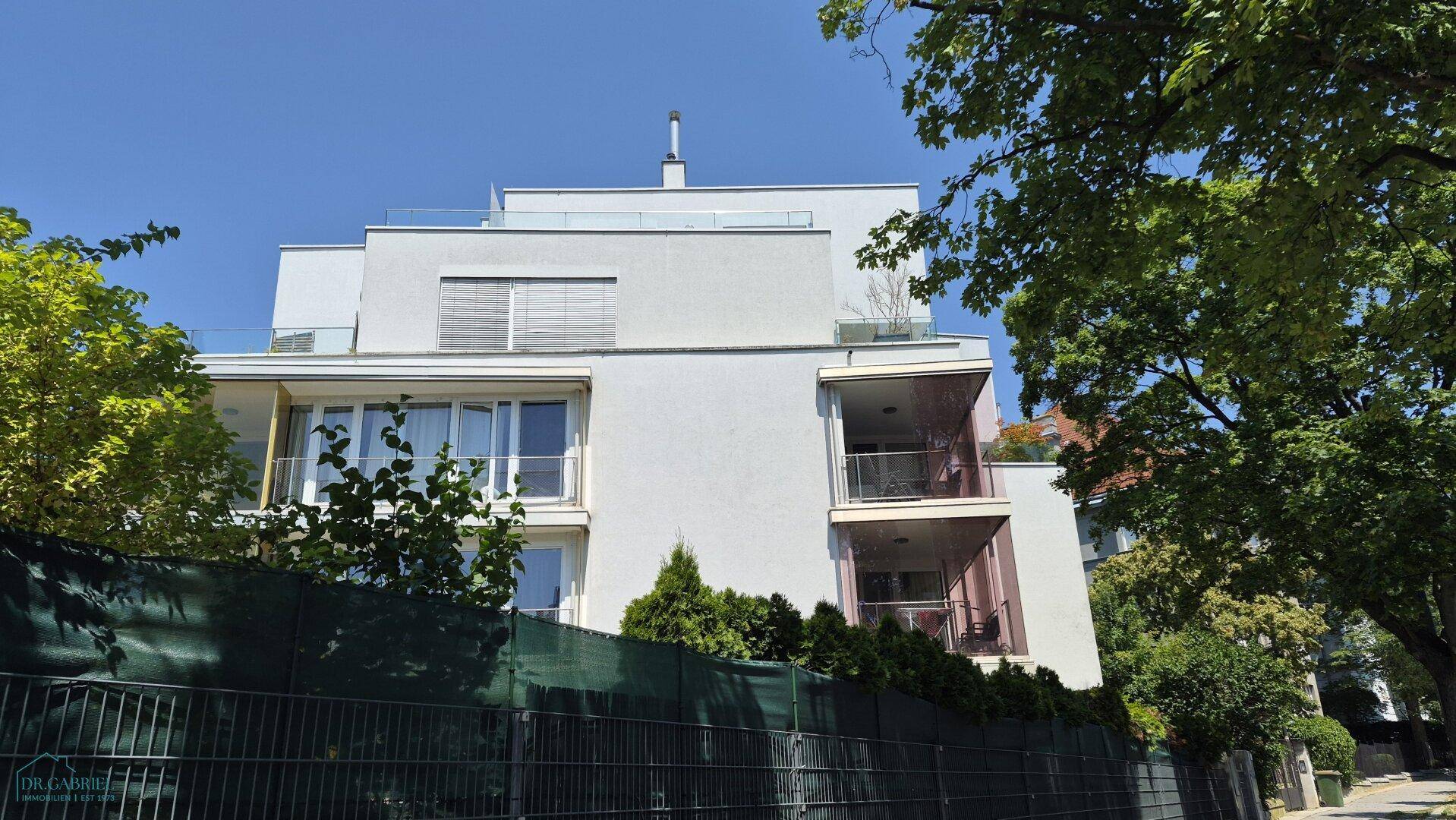 Moderne Wohnhausfassade mit Balkonen und großen Fenstern, umgeben von Bäumen.