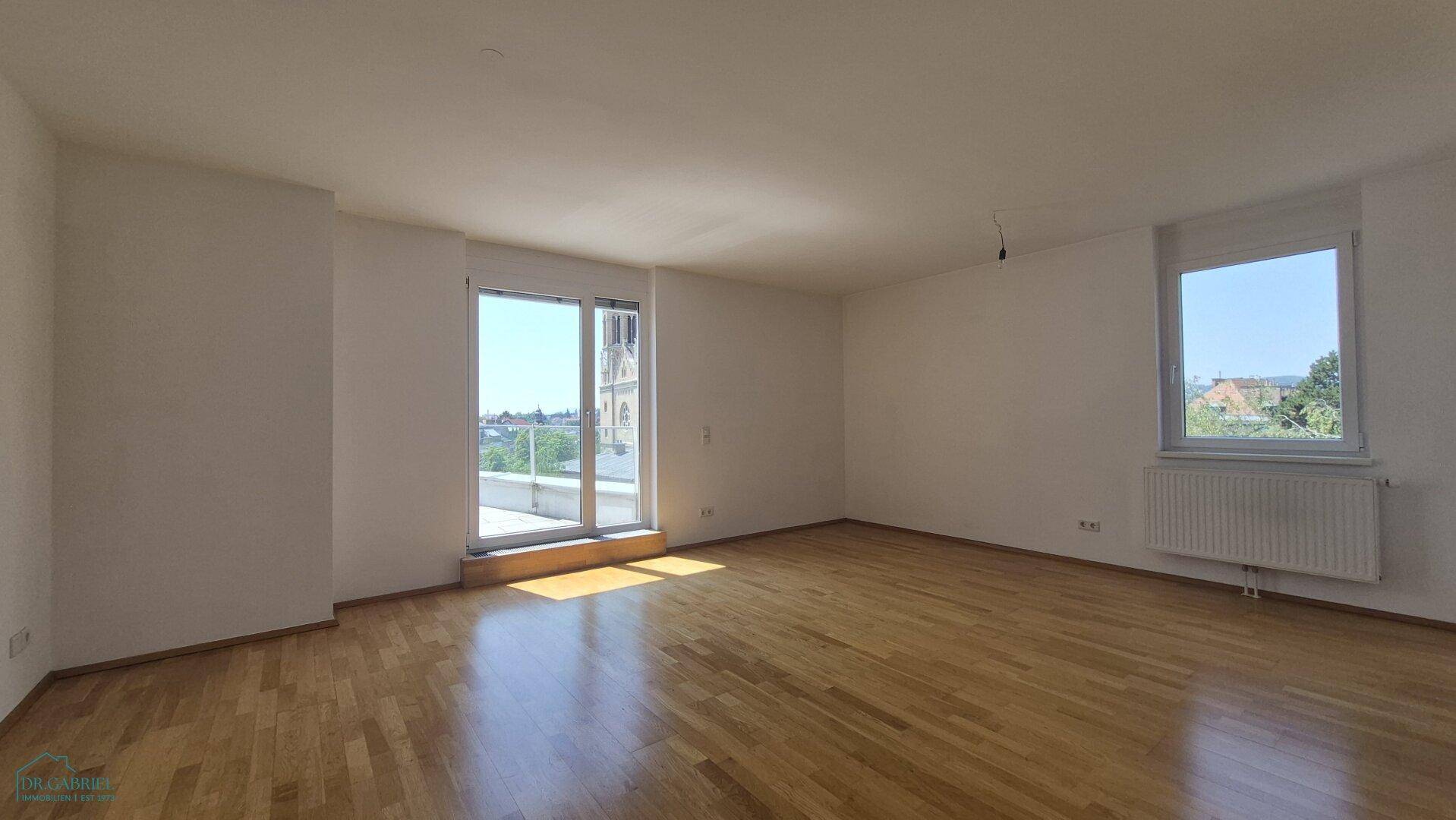 Geräumiges, leeres Wohnzimmer mit Holzboden, großen Fenstern und viel Tageslicht.
