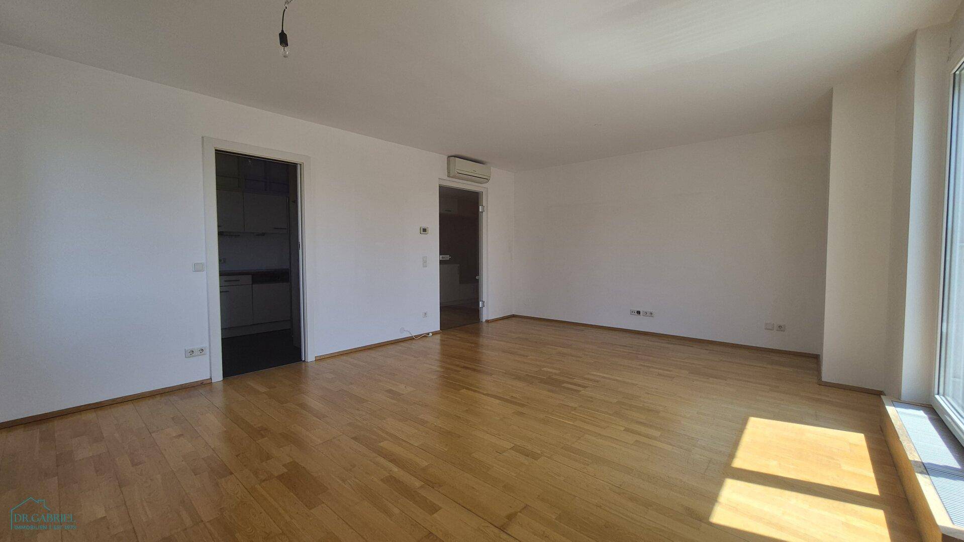 Leeres Wohnzimmer mit Holzboden, Klimaanlage und Zugang zu weiteren Räumen.