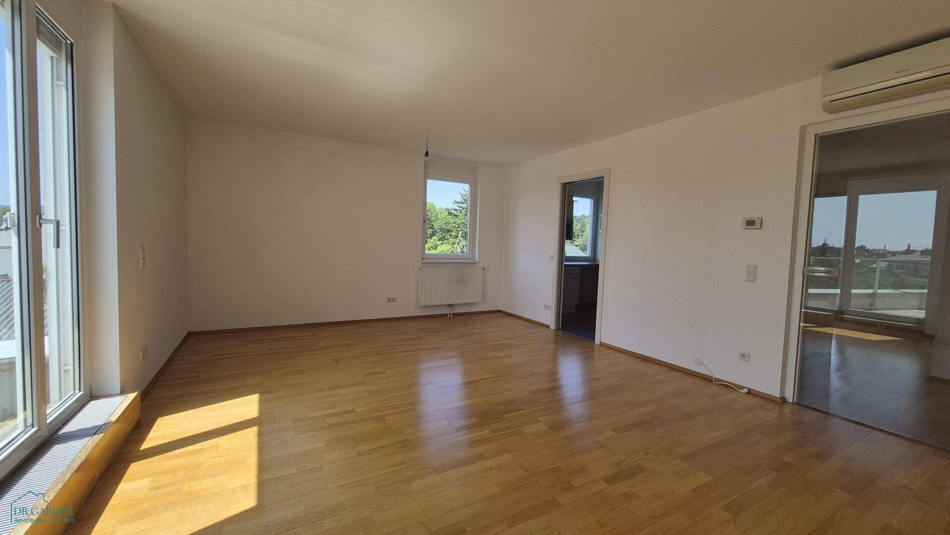 Geräumiges Wohnzimmer mit Holzboden, großen Fenstern und Klimaanlage.