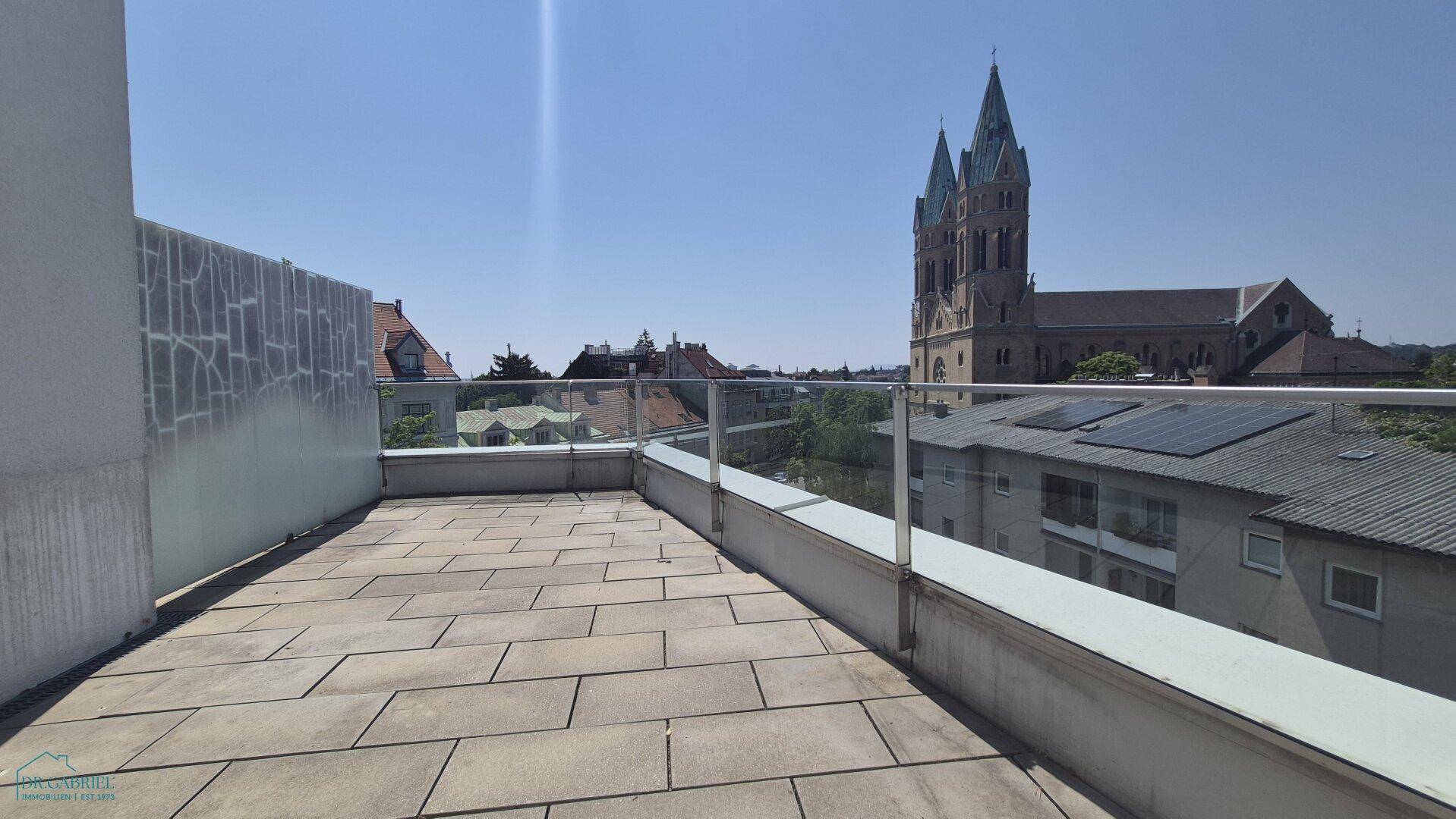 Unmöblierte Terrasse mit Steinplatten, Glasgeländer und weitem Blick über die Umgebung.