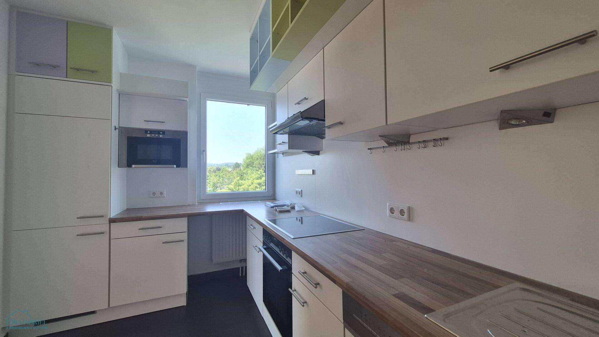 Moderne Einbauküche mit weißen Schränken, Holzarbeitsplatte und Fenster mit Grünblick.
