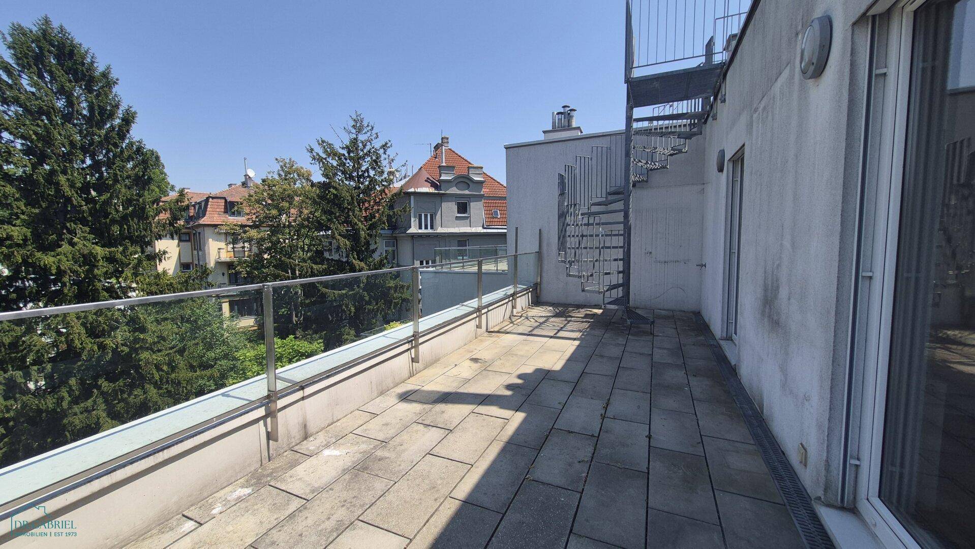 Große Terrasse mit Steinplatten, Glasgeländer und einer Außentreppe.