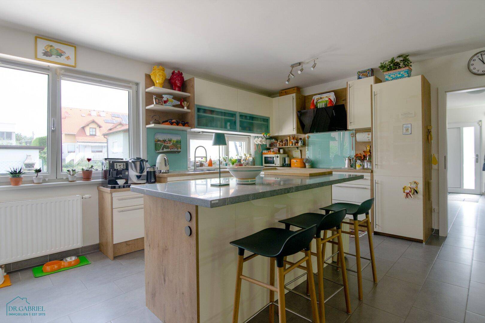 Moderne Küche mit Kücheninsel, hellen Schränken und Fliesenboden, mit Blick nach draußen.