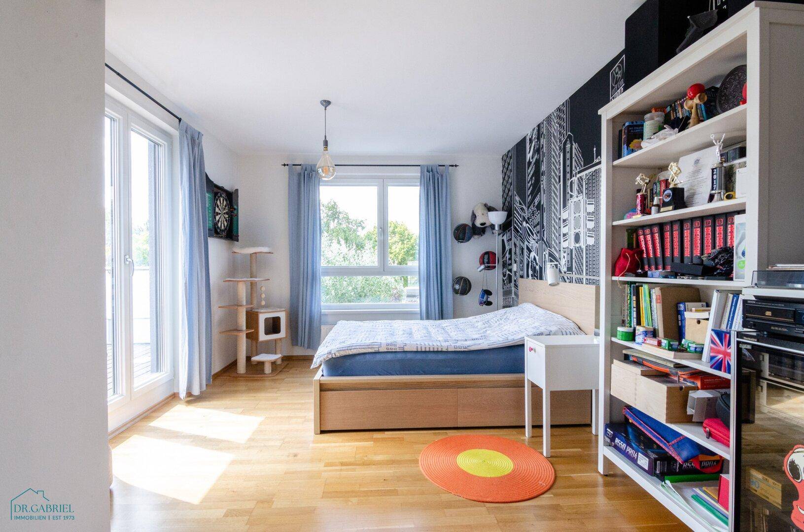Helles Kinder- oder Gästezimmer mit Bett, Bücherregal und Zugang zum Balkon.