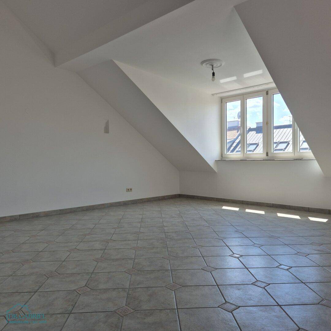 Heller Raum mit schrägen Wänden, Fliesenboden und einem großen Fenster, das viel Tageslicht hereinlässt.