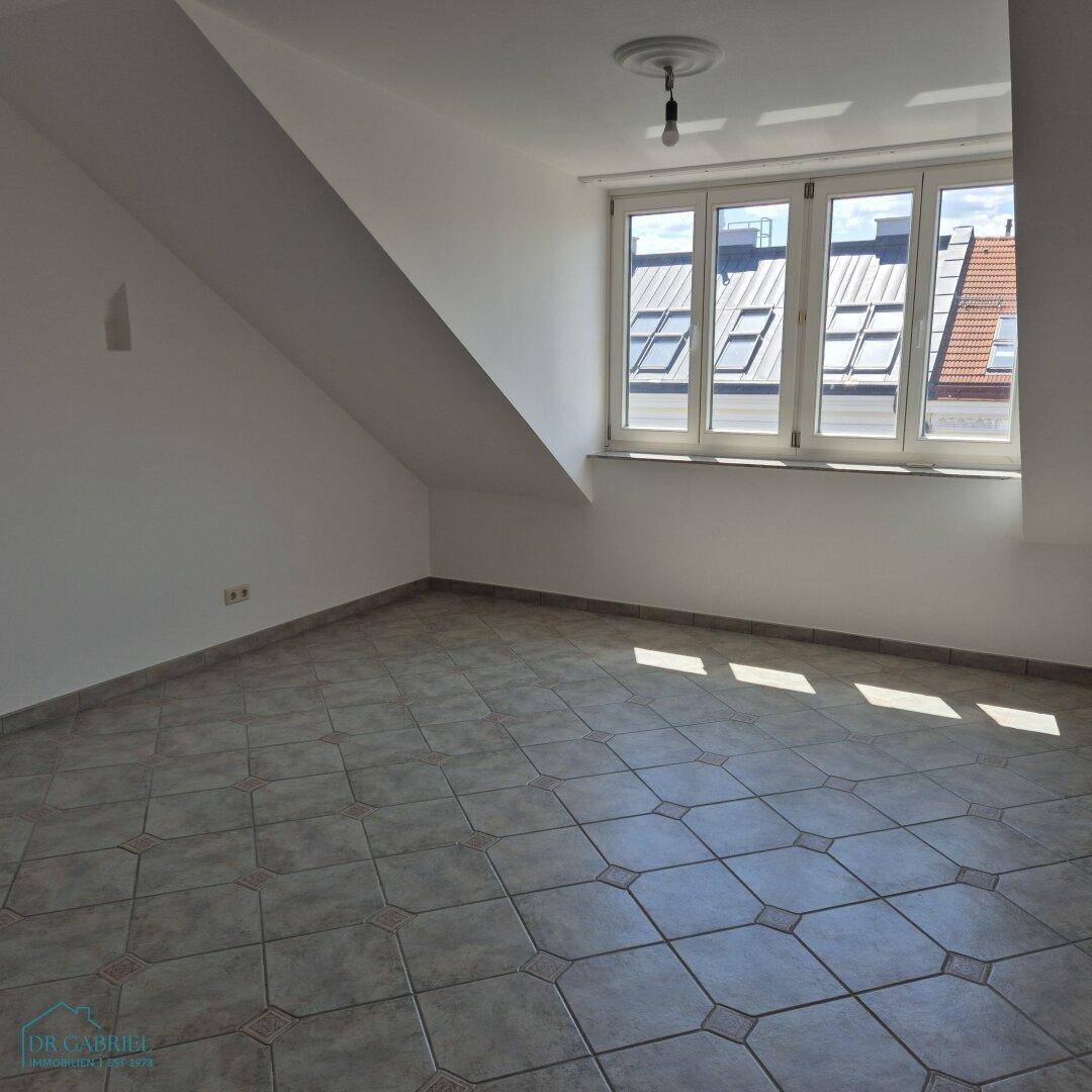 Geräumiger Raum mit Fliesenboden, schrägen Wänden und großen Fenstern, die den Raum mit Licht füllen.