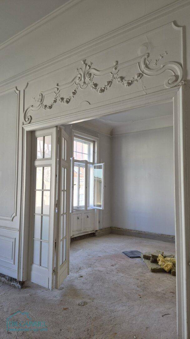 Geräumiger Innenraum in Renovierung mit kunstvollen Stuckelementen über der Tür und Blick durch große Fenster.