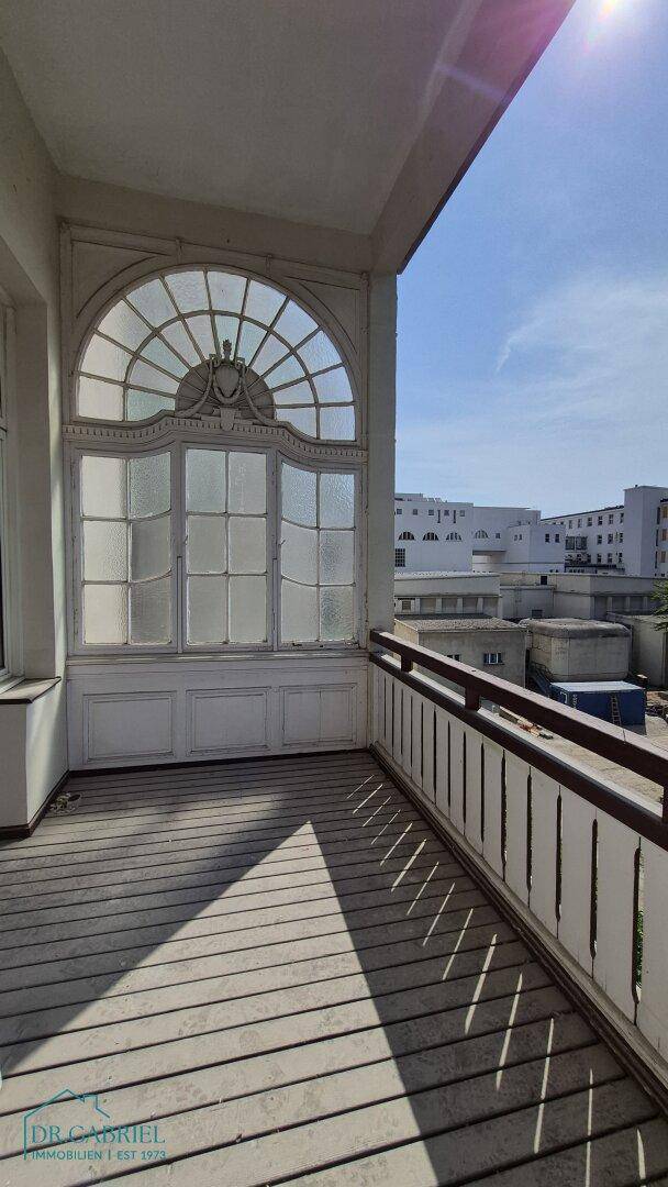 Sonniger Balkon mit Holzdielen und kunstvollem Bogenfenster, bietet einen weiten Blick über die Stadt.