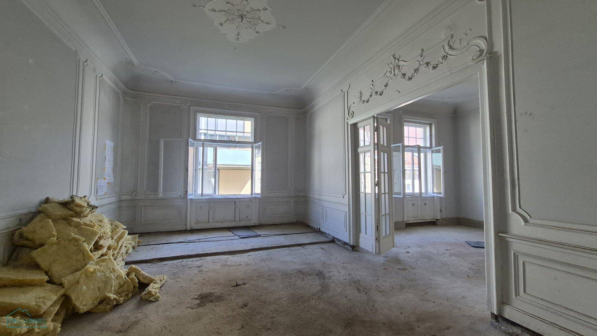 Großer Raum in Renovierung mit kunstvollen Stuckverzierungen an Wänden und Decke sowie großen Fenstern.