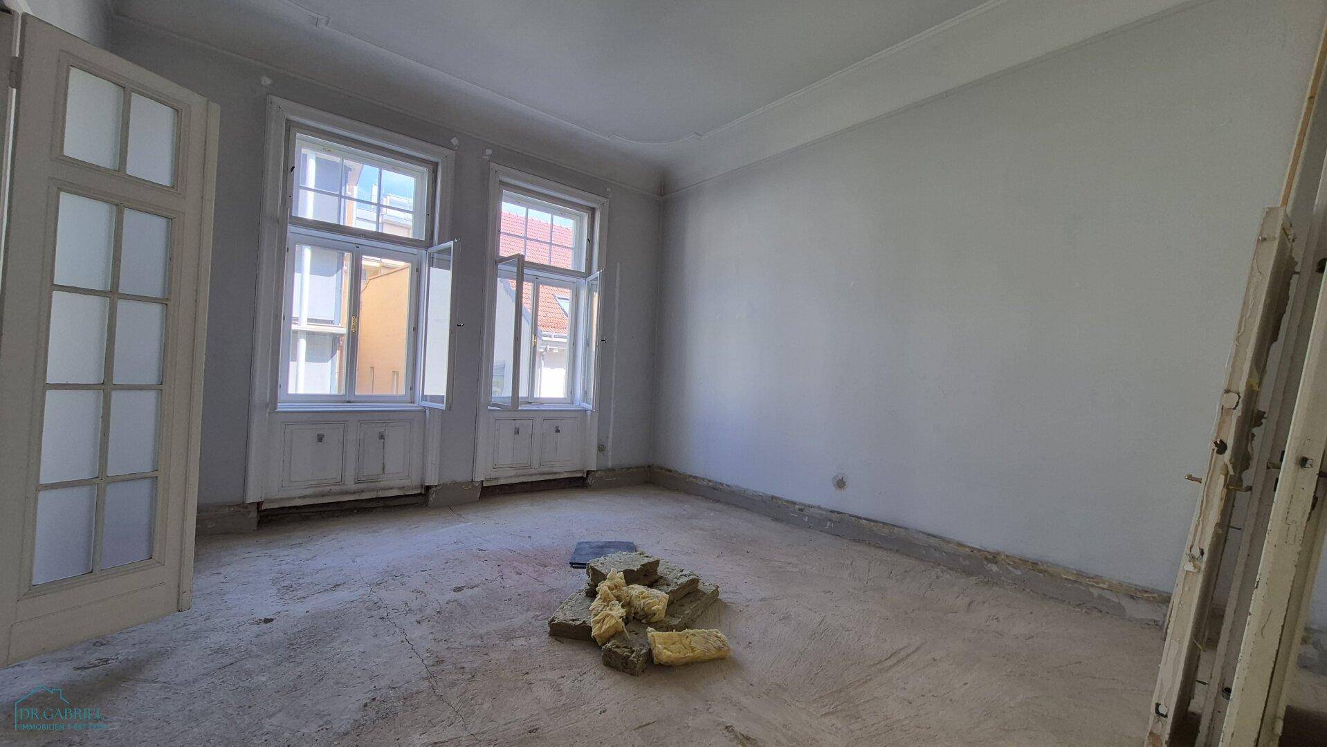 Heller Raum in Renovierung mit Betonboden und zwei Fenstern, die viel Tageslicht hereinlassen.