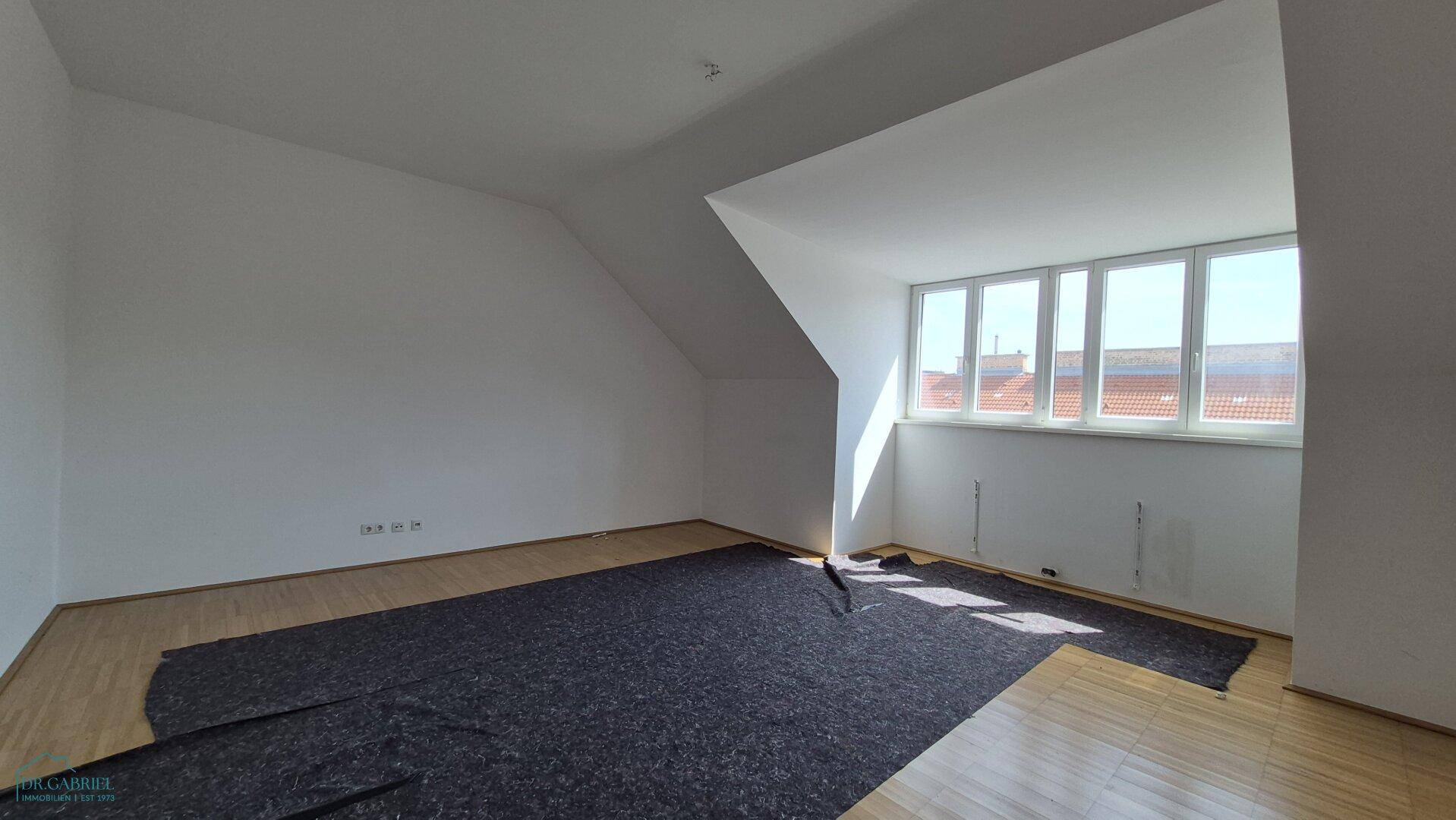 Geräumiges, leeres Zimmer mit Parkettboden, Dachschrägen und breiter Fensterfront.