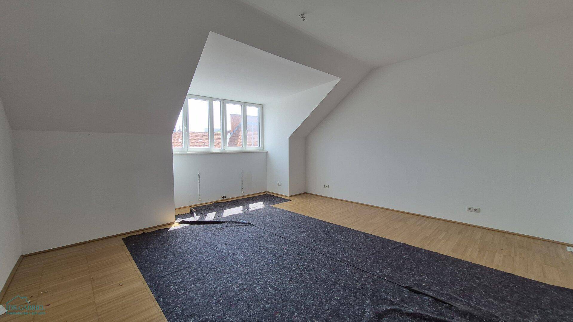 Helles, leeres Zimmer mit Parkettboden, Dachschrägen und viel natürlichem Lichteinfall.