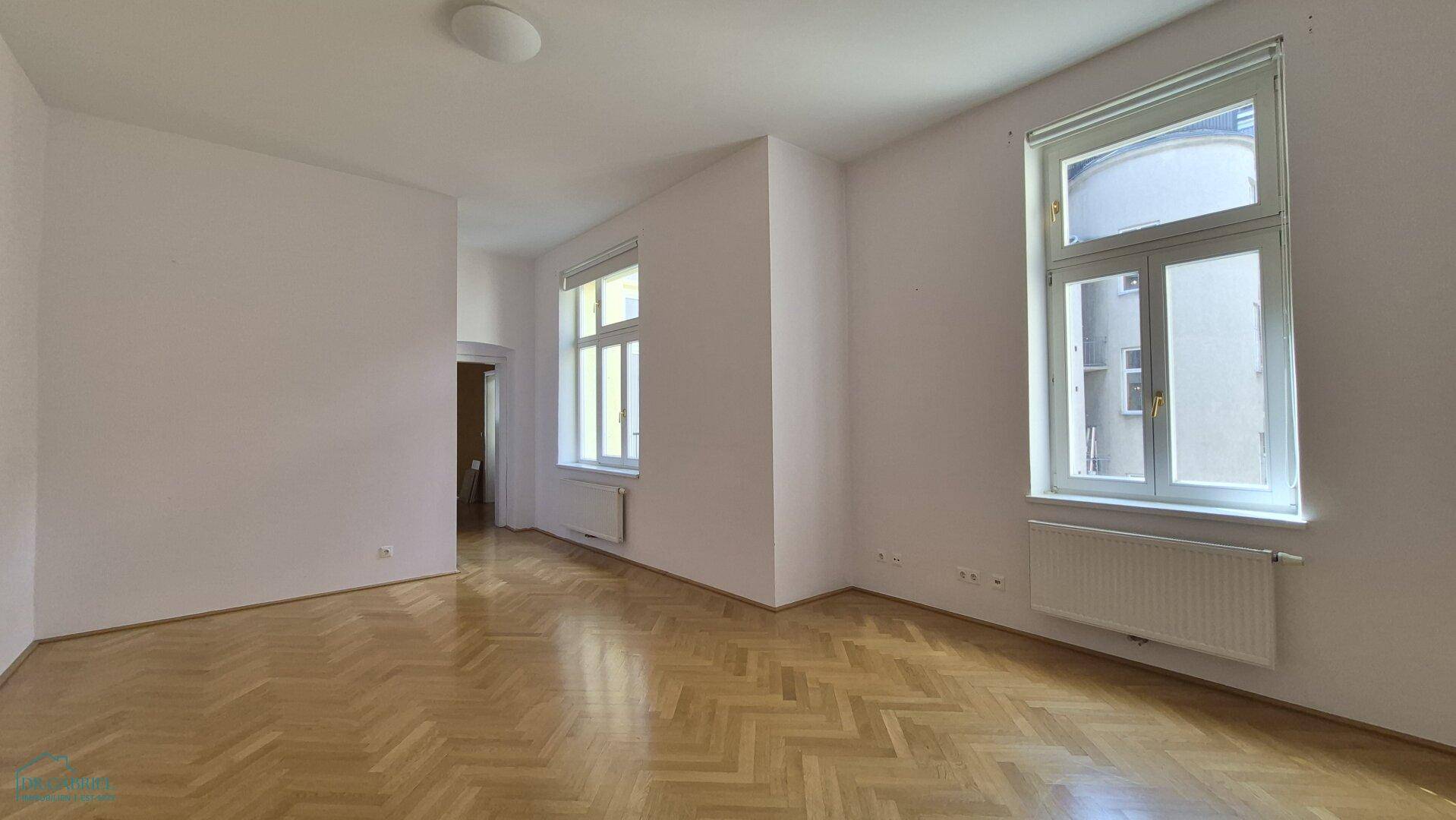 Lichtdurchflutetes Zimmer mit Fischgrätparkett und hohen Fenstern, bereit zur individuellen Gestaltung.