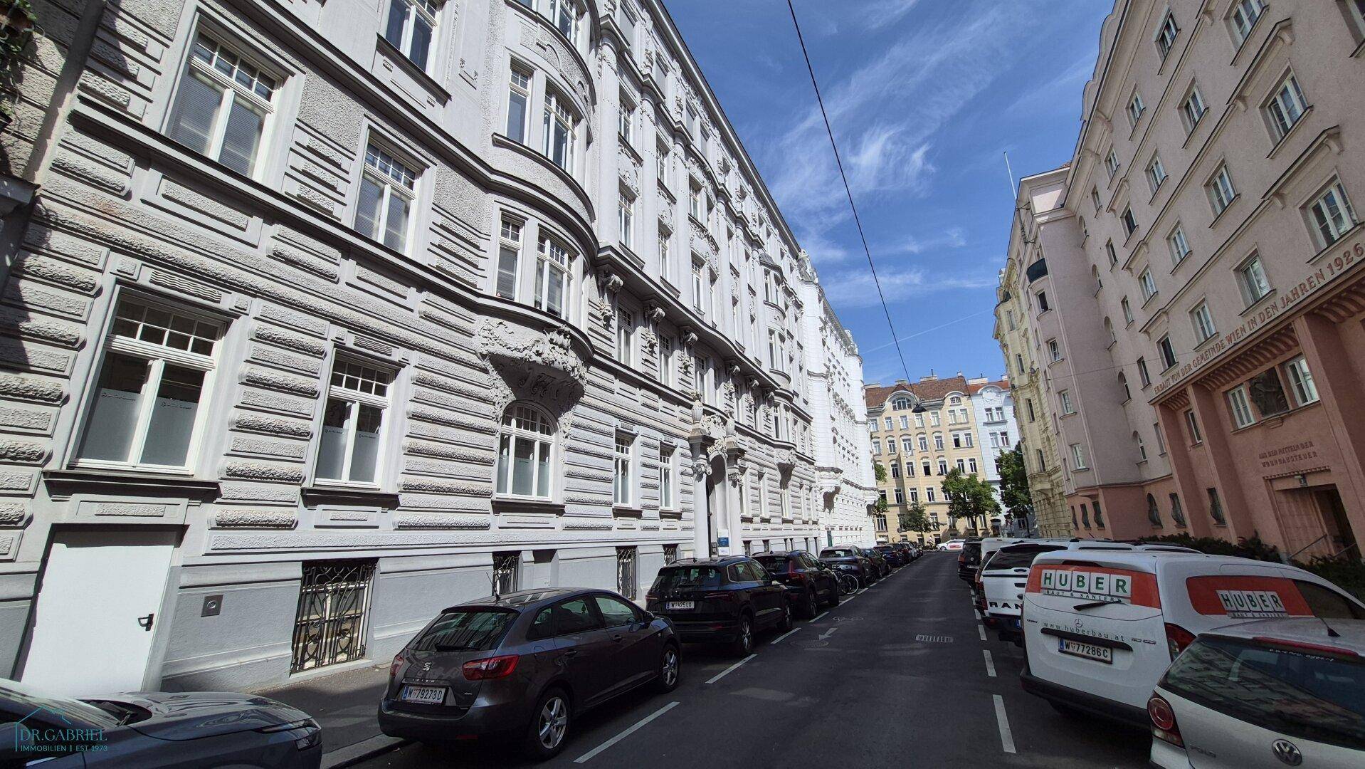 Historische Straßenansicht mit Altbaufassaden und parkenden Autos an einem sonnigen Tag.