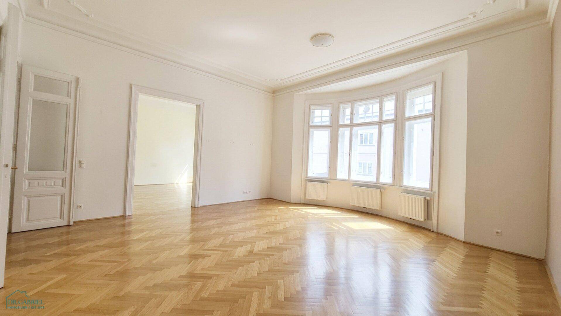 Helles Wohnzimmer mit Fischgrätparkett, Stuckdecken und Zugang zu weiteren Räumen.
