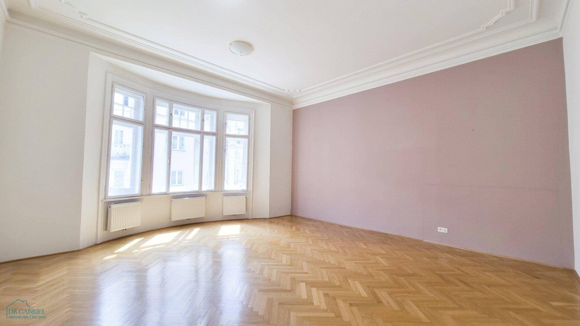 Geräumiges, leeres Wohnzimmer mit hellem Fischgrätparkett, Stuckdecken und einem großen Erkerfenster.
