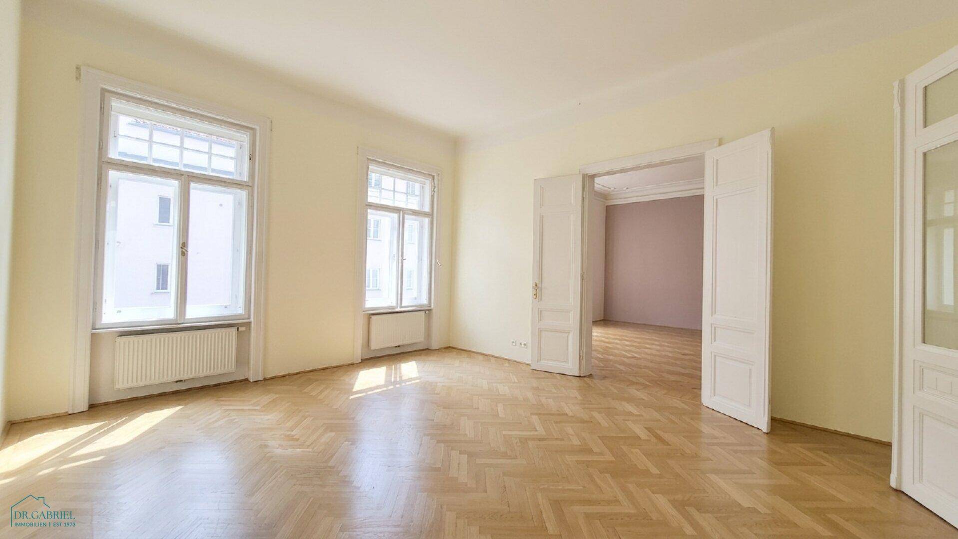 Geräumiges Zimmer mit Fischgrätparkett und hohen Fenstern, das viel Tageslicht hereinlässt.