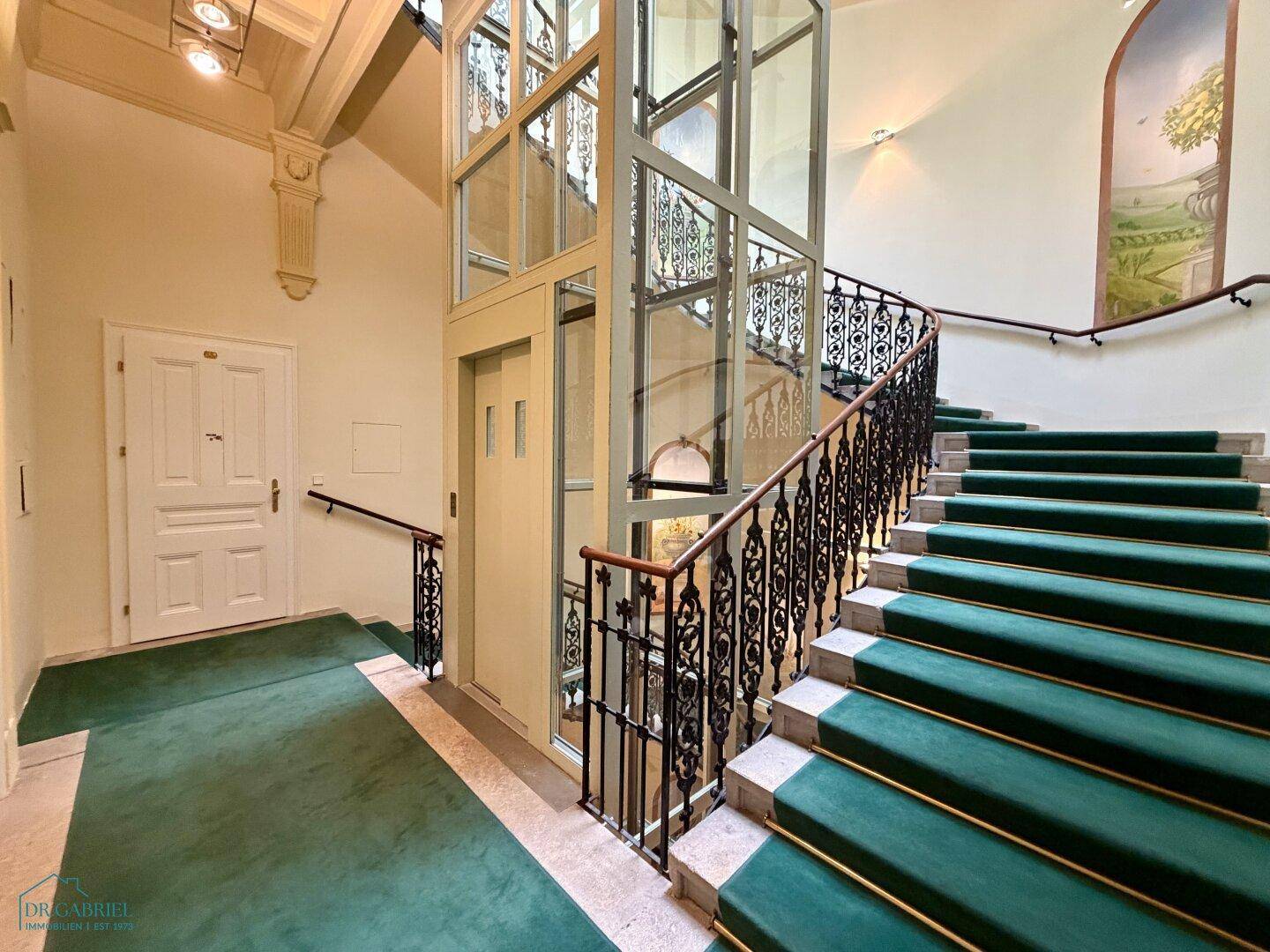Elegantes Treppenhaus mit Teppich, schmiedeeisernem Geländer und einem modernen Glasaufzug.