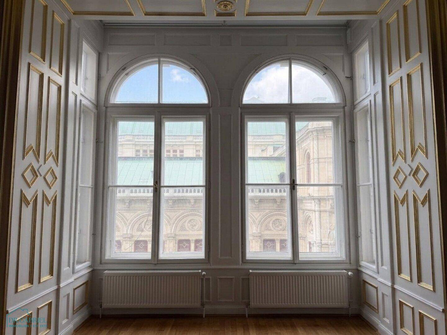 Ein eleganter Raum mit hohen Fenstern, Stuckverzierungen und Blick auf ein historisches Gebäude.