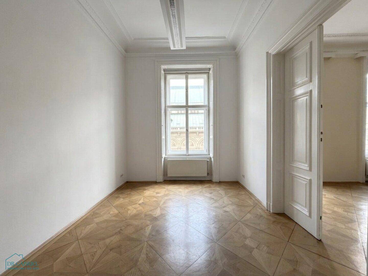 Heller Raum mit Parkettboden, hohen Decken und einem großen Fenster mit Blick nach draußen.