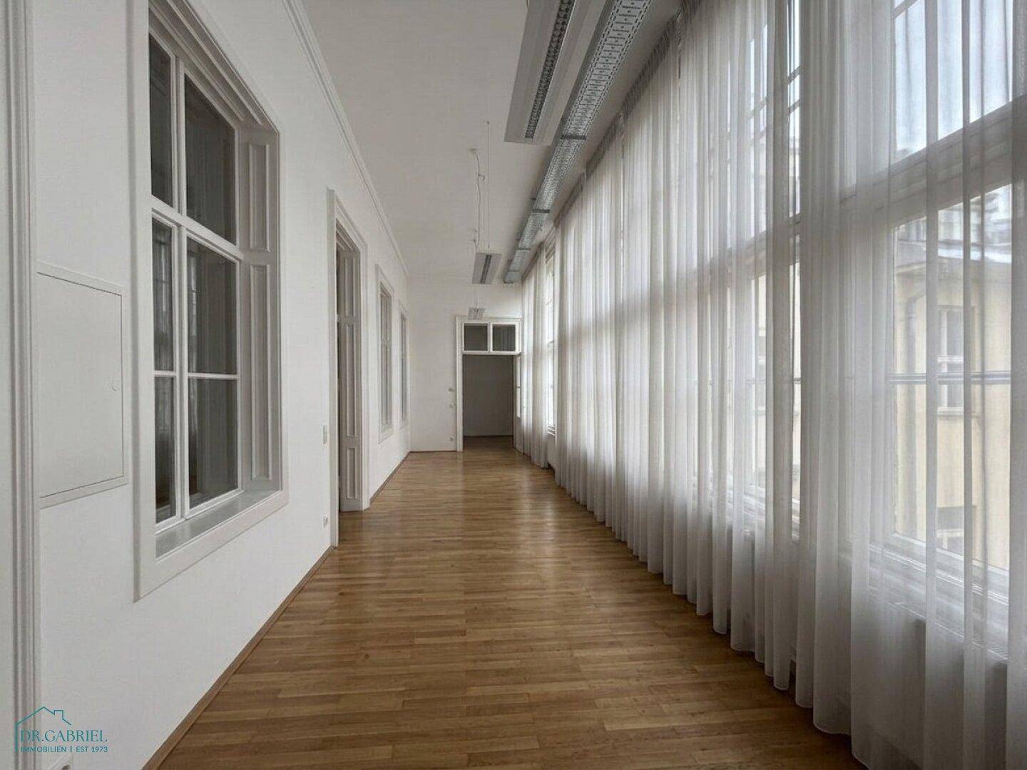 Langer Flur mit Holzboden, hohen Fenstern und Vorhängen, der zu weiteren Räumen führt.