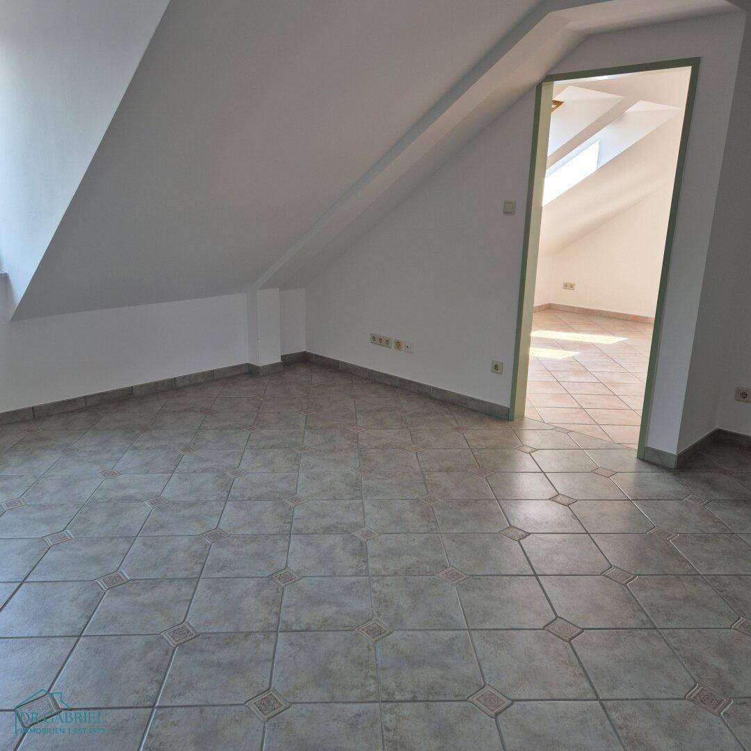 Leerer Raum im Dachgeschoss mit schrägen Wänden und Fliesenboden, hell durch Dachfenster.