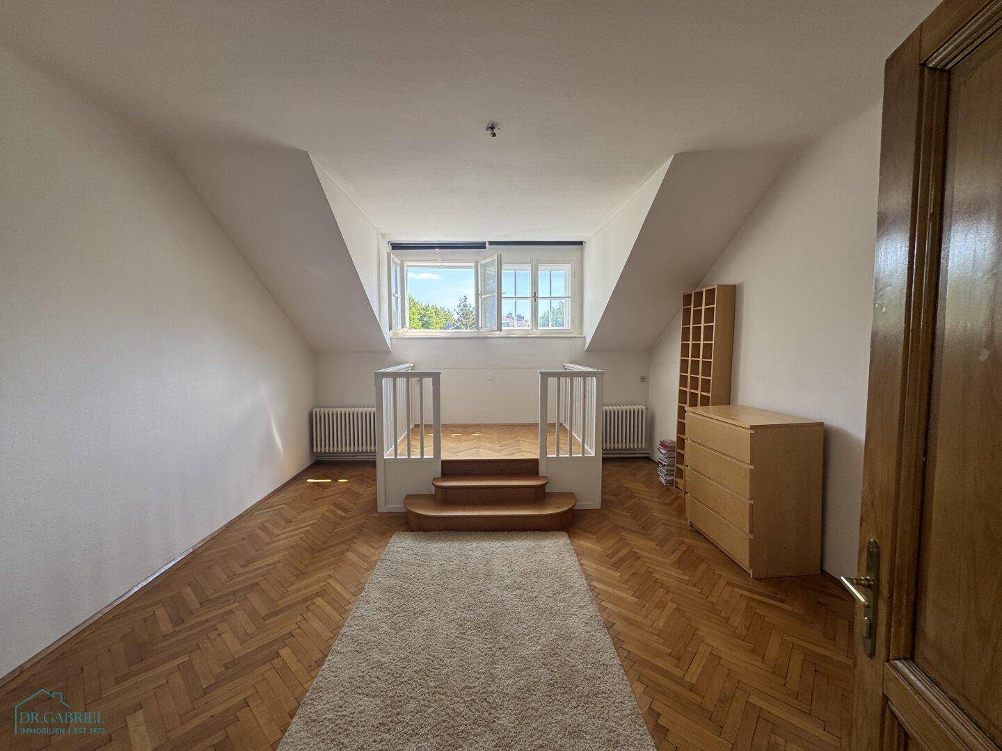 Dachzimmer mit schrägen Wänden, Fischgrätparkett und einem erhöhten Bereich mit Fenster.