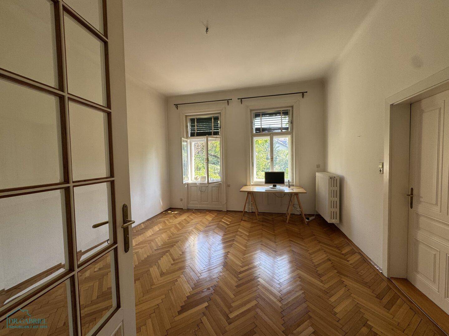 Helles Arbeitszimmer mit Fischgrätparkett, großen Fenstern und einem einfachen Schreibtisch.