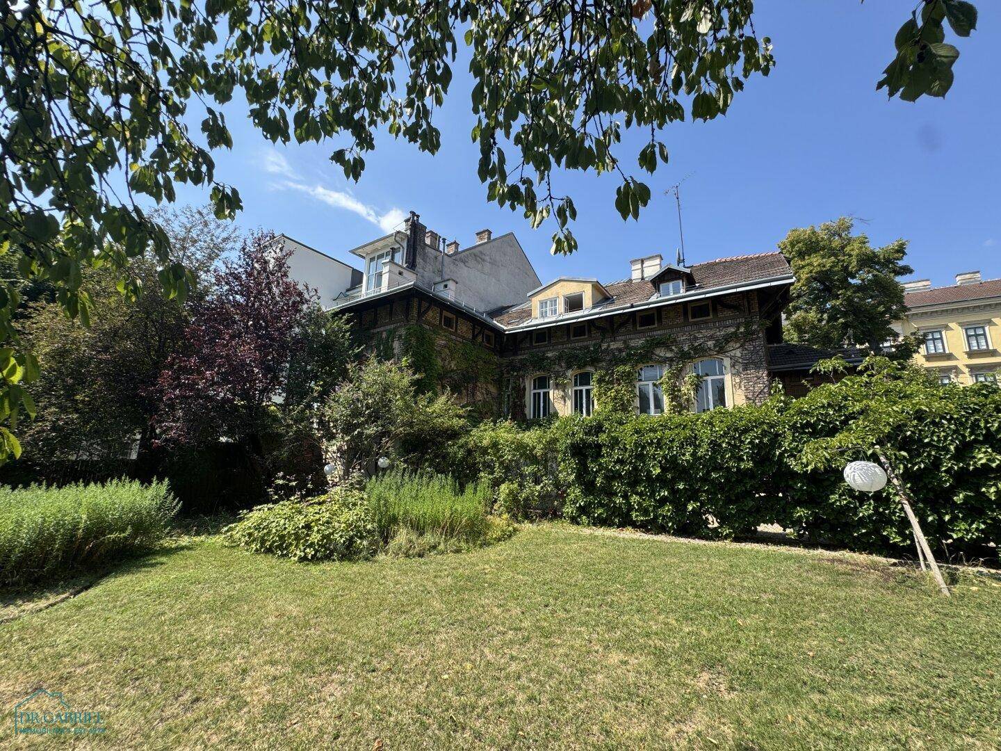 Historisches Gebäude, umgeben von einem großen, grünen Garten mit Hecken und Bäumen.