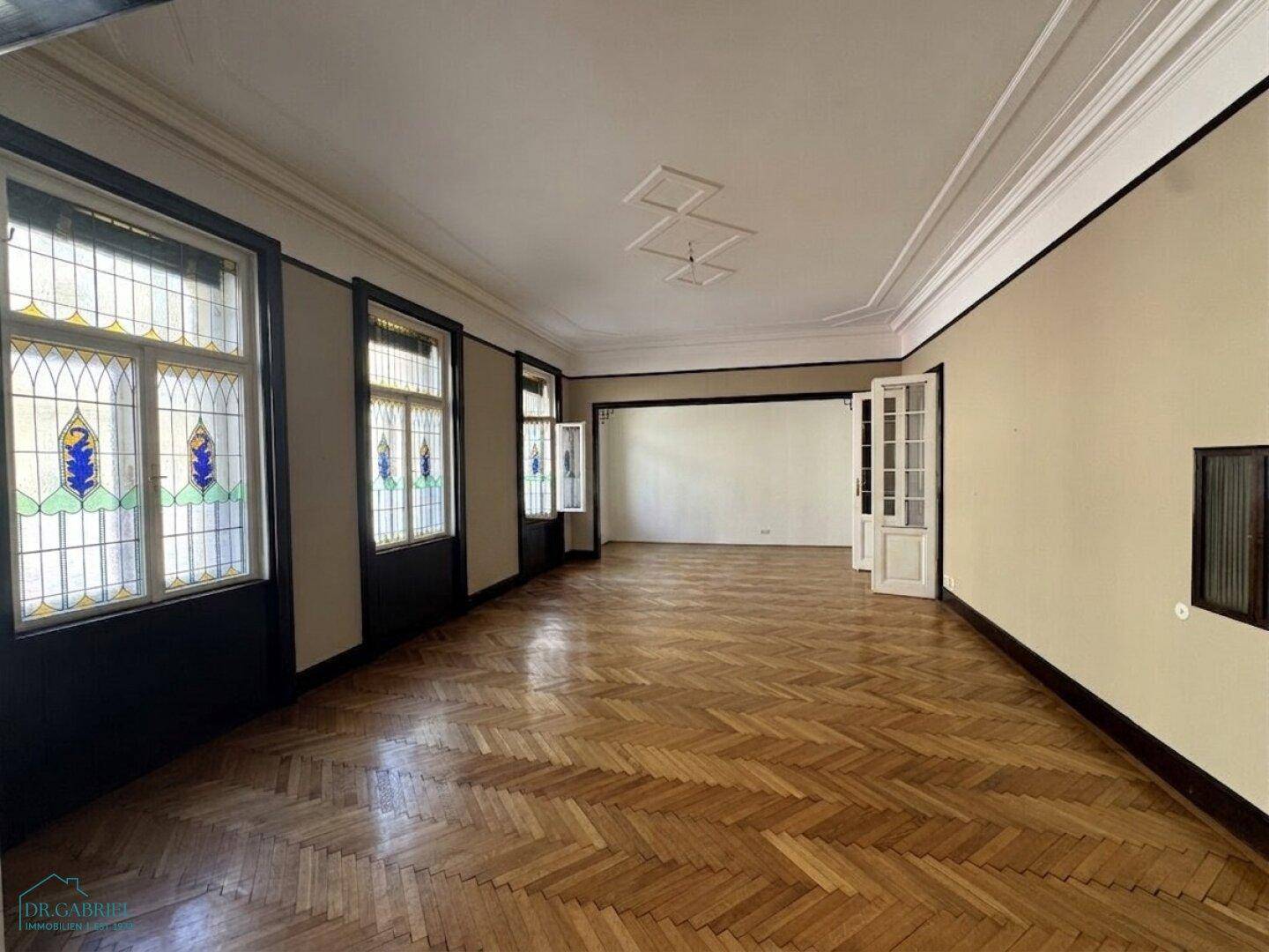 Geräumiges Wohnzimmer mit Fischgrätparkett, hohen Decken und dekorativen Buntglasfenstern.
