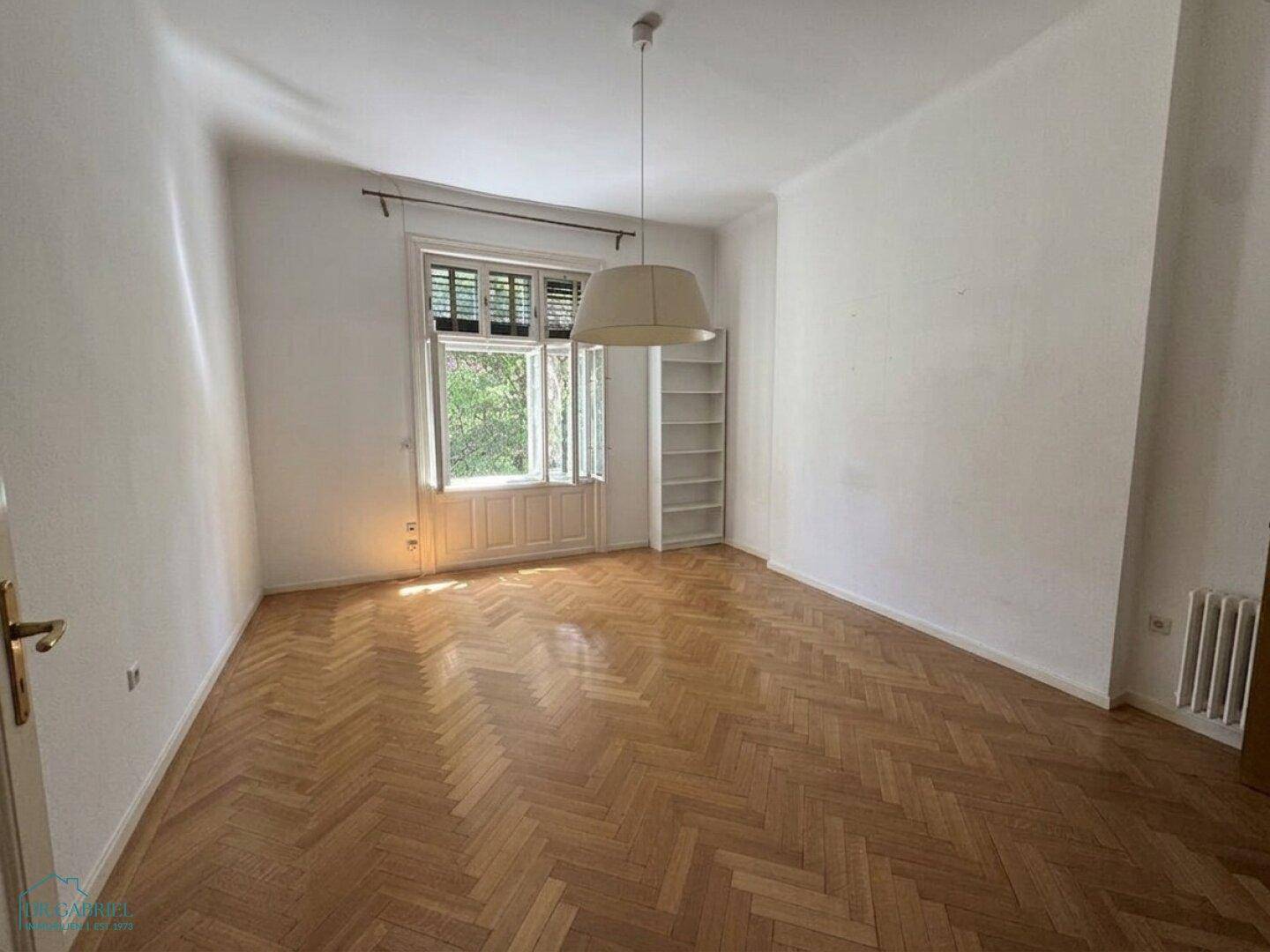 Helles Zimmer mit Fischgrätparkett, einem Fenster und einem einfachen Bücherregal.
