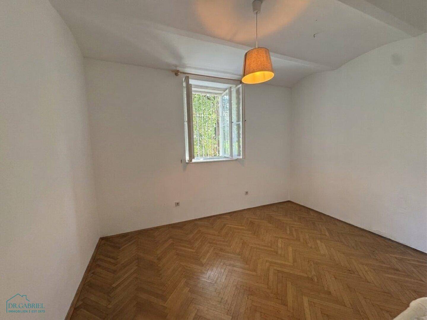 Helles Zimmer mit Fischgrätparkett und einem geöffneten Fenster, das viel Licht hereinlässt.