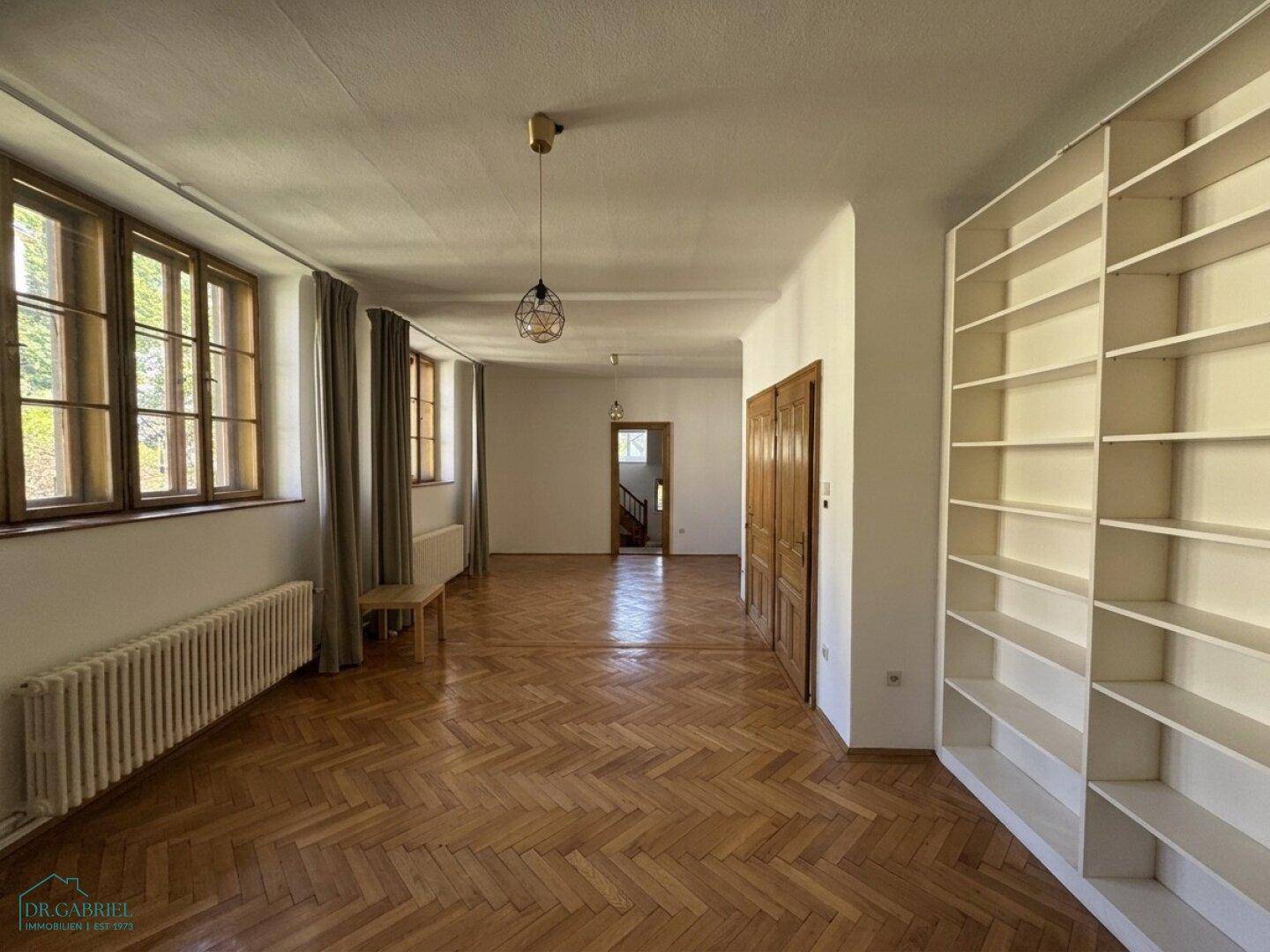 Geräumiger Wohnbereich mit Fischgrätparkett, hohen Fenstern und einem großen Bücherregal.