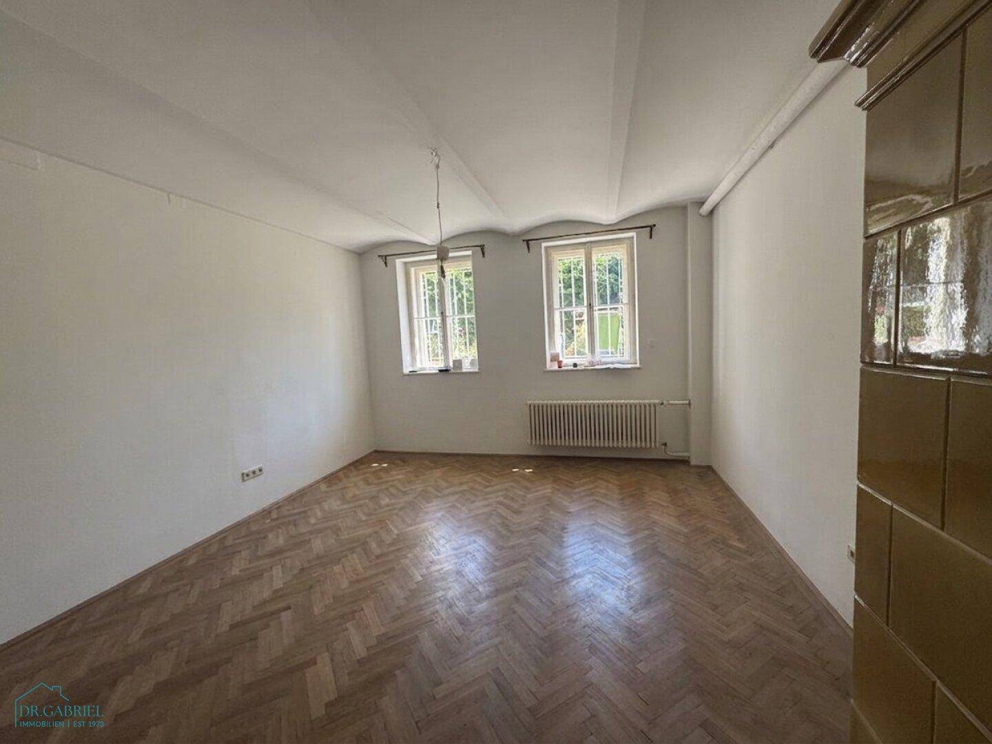 Geräumiges Zimmer mit Fischgrätparkett, zwei Fenstern und einem traditionellen Kachelofen.