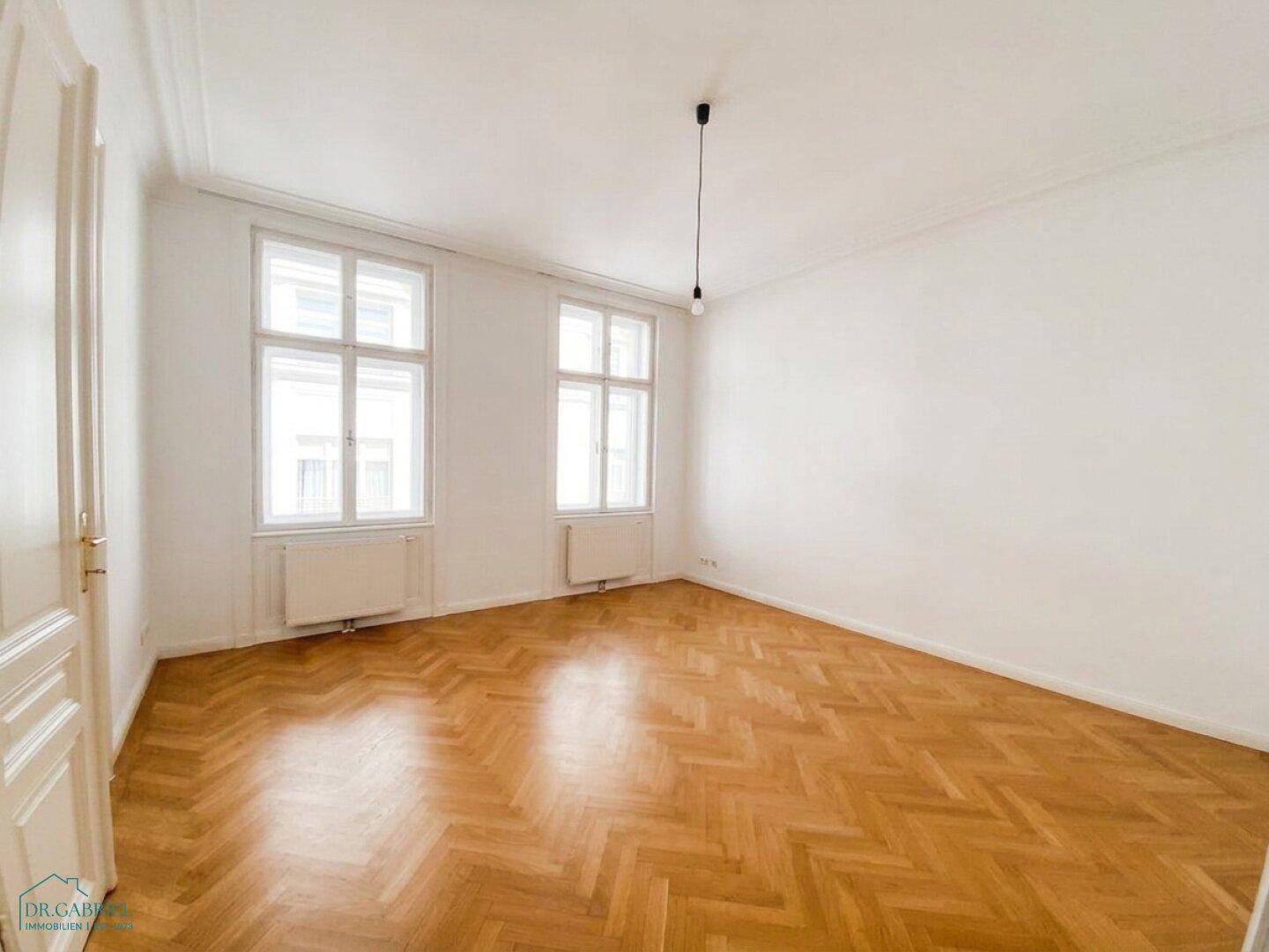 Lichtdurchflutetes Zimmer mit klassischem Fischgrätparkett und zwei großen Fenstern.