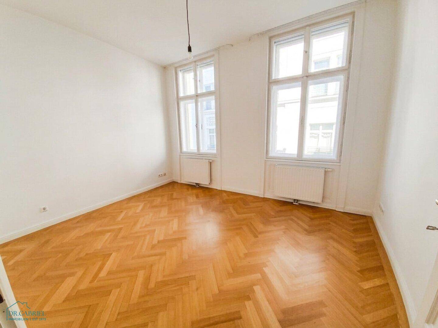 Leeres Zimmer mit Fischgrätparkett und zwei großen Fenstern, ideal für individuelle Einrichtung.