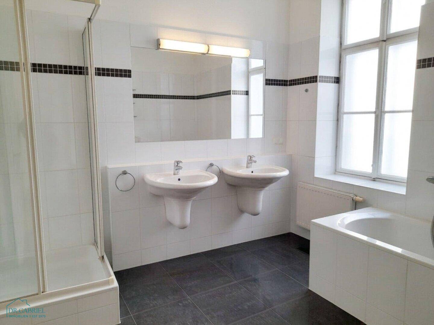 Helles Badezimmer mit zwei Waschbecken, Badewanne und Dusche, gut beleuchtet durch ein großes Fenster.
